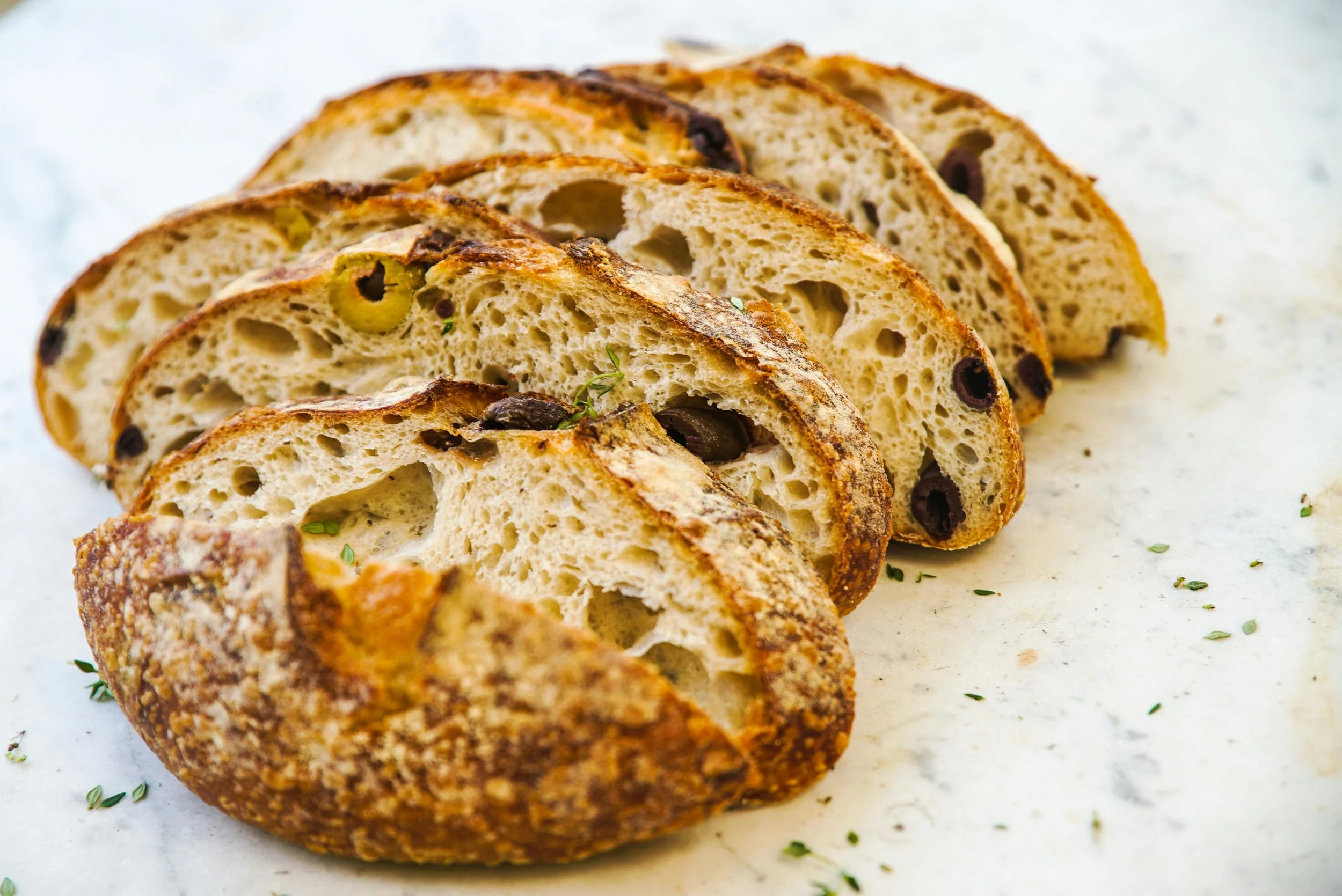 Tranches de pain artisanal aux olives, à la croûte dorée, disposées sur une surface blanche.