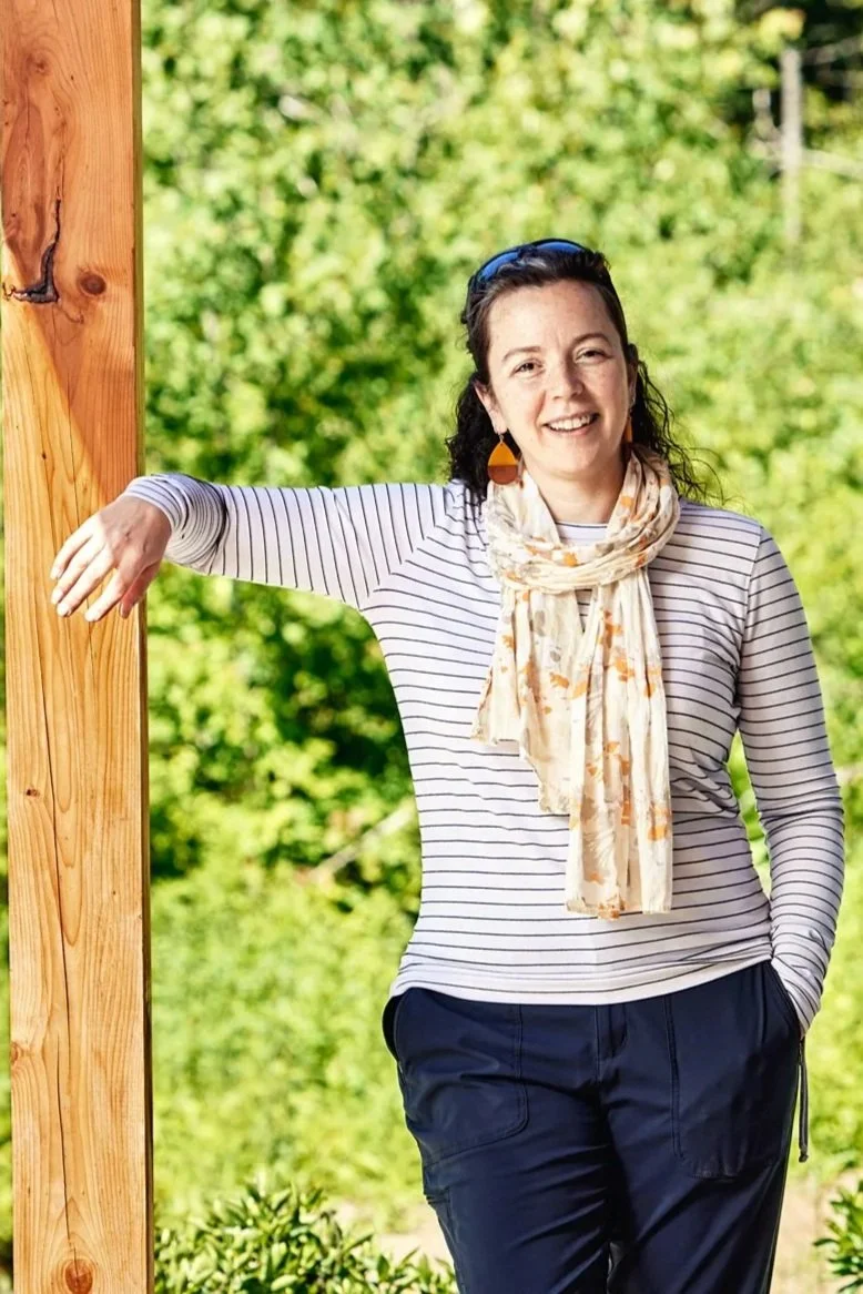 Photo of Emily Mottram smiling on front porch leaning on beam wearing a scarf and striped shirt