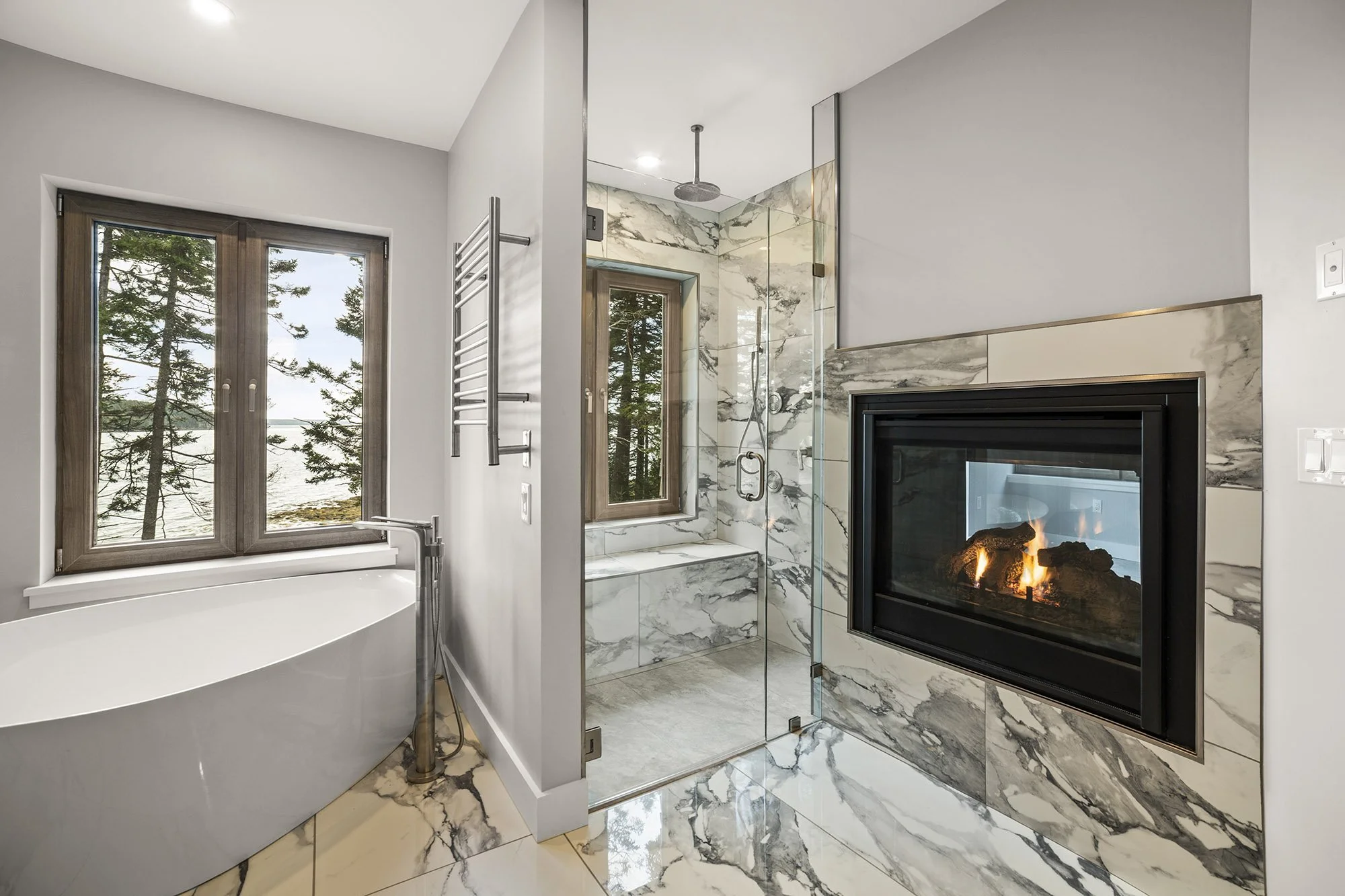 Gorgeous 5 Piece Master Bathroom with a fireplace that can be seen in the other room, a tiled shower with a waterfall rain showerhead, a window that looks out a bench in teh shower, and a deep soaker tub with a window that overlooks the water