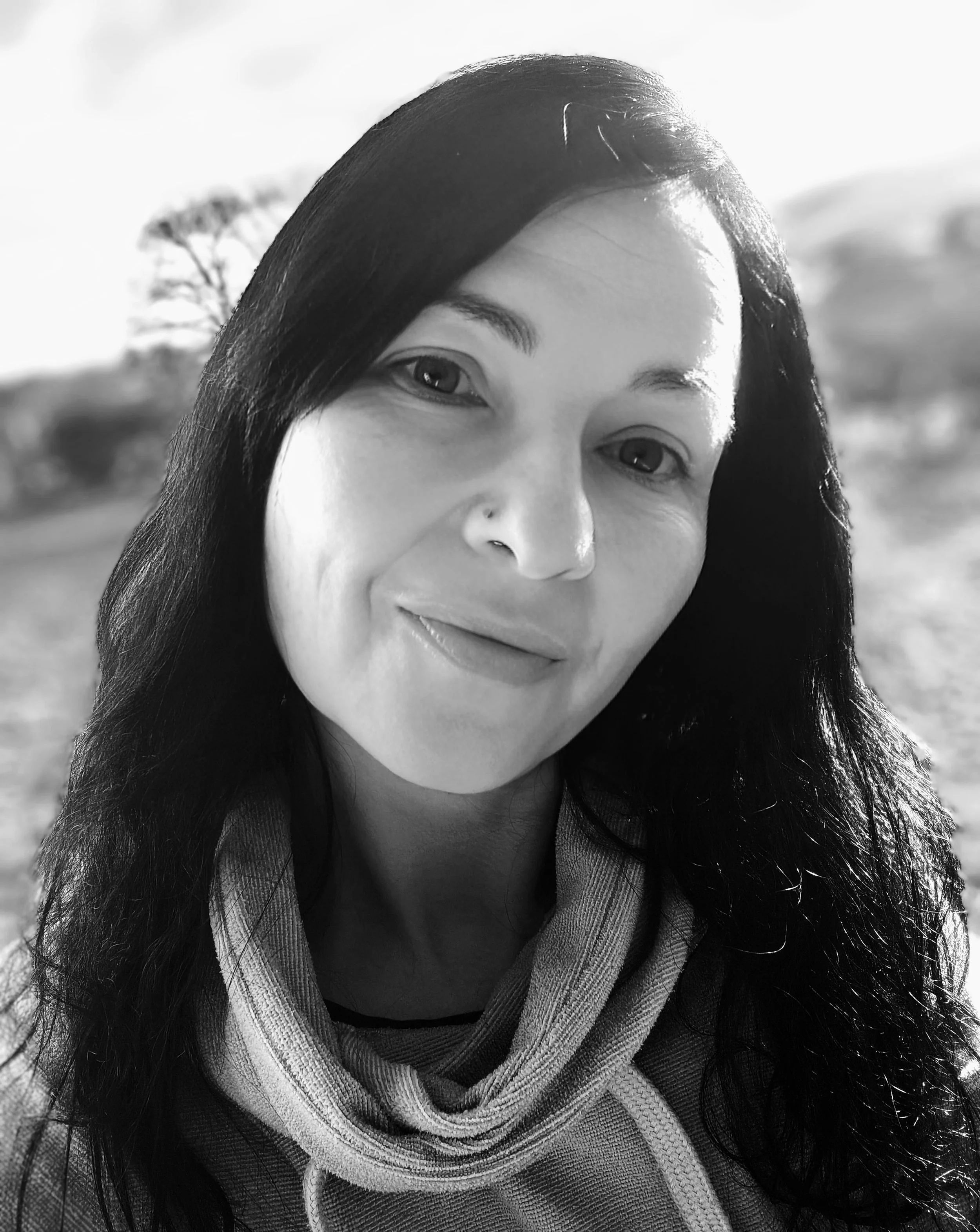 Black and white portrait of the woman behind Spirit Soul Bliss with long dark hair, smiling outdoors with a blurred background of trees and sky.