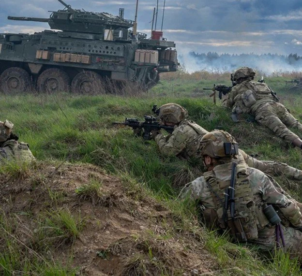 Military soldiers in camouflage uniforms lying prone in a grassy field, aiming rifles towards a tank in the distance.
