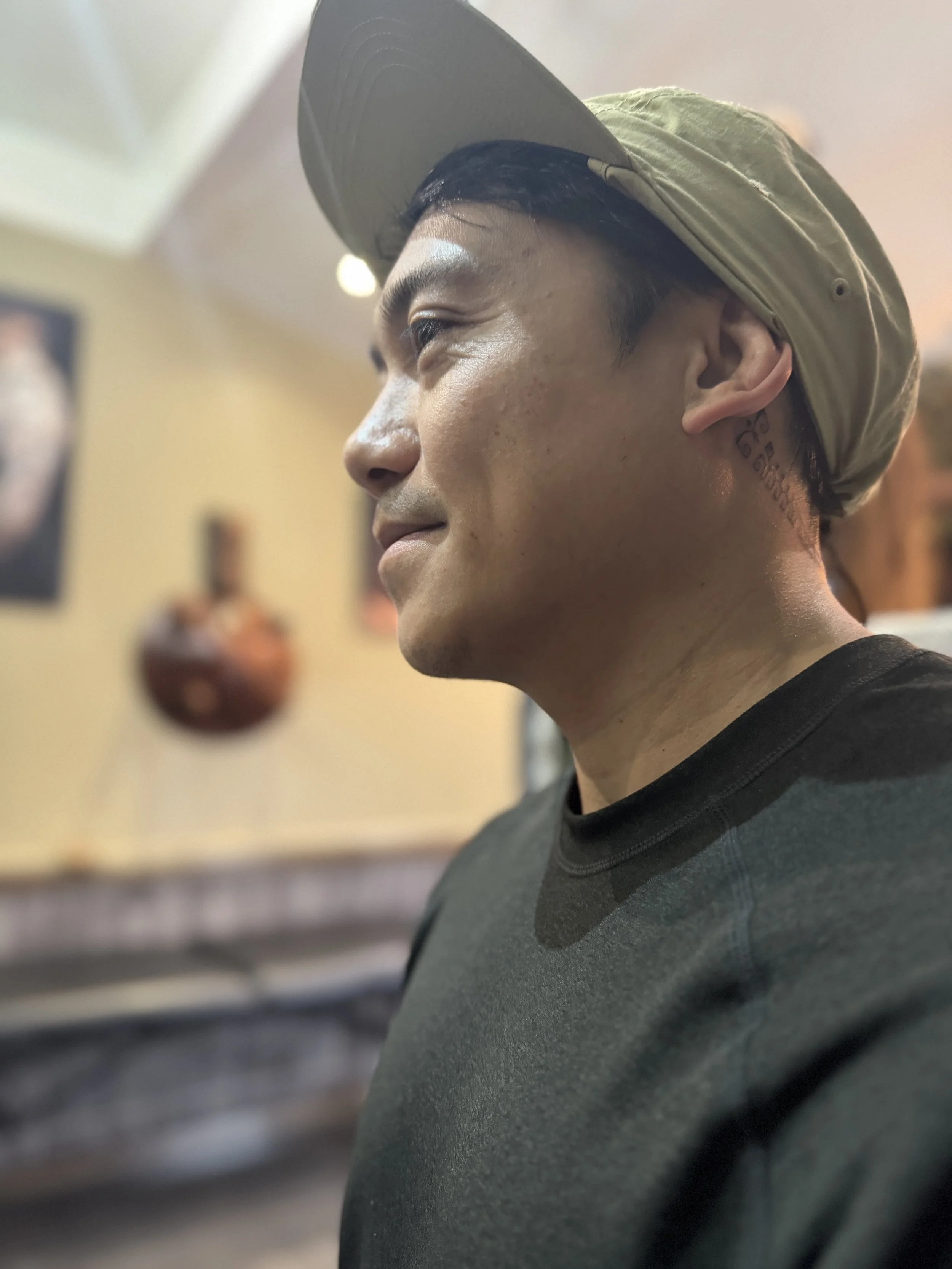 Profile of a young man wearing a gray cap and a black shirt, smiling softly with a tattoo visible behind his ear, indoors with blurred background.