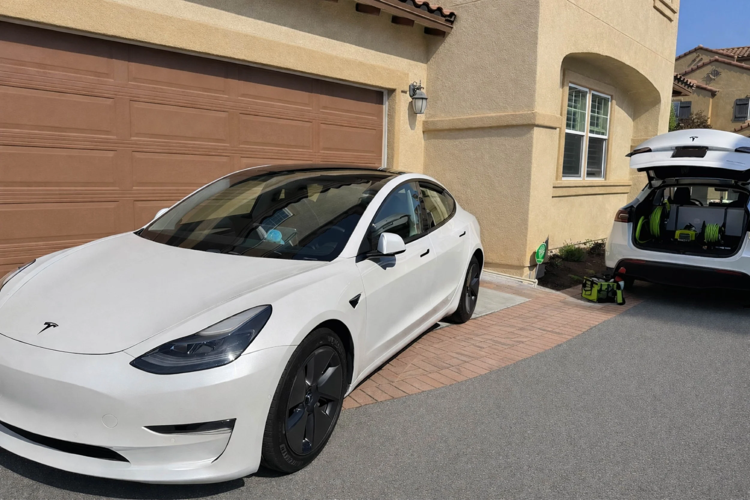 White Tesla Model 3 parked in front of a house with a garage door, with the mobile detailing car trunk open containing green hoses and equipment. A toolbox and a small green sign are on the ground nearby.