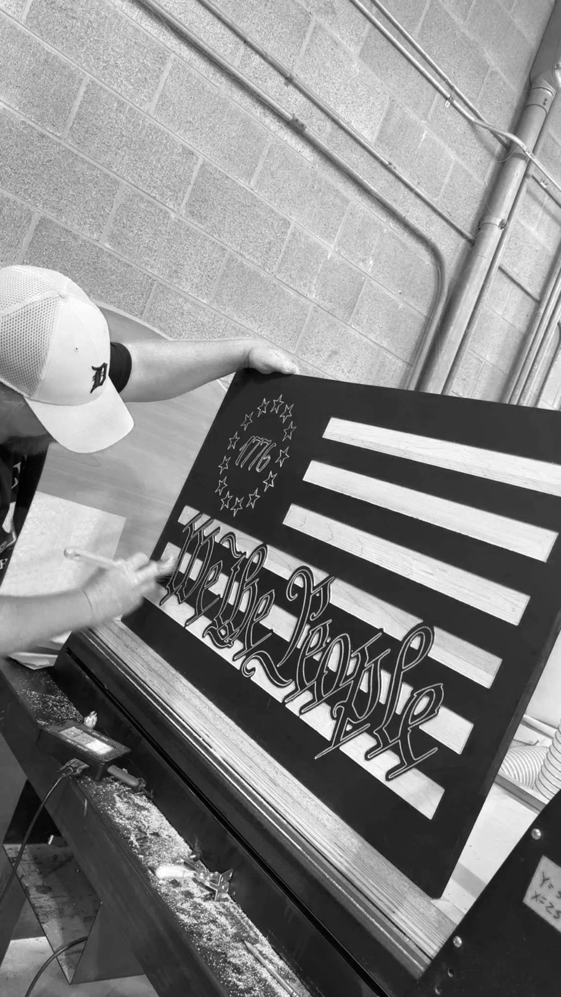 Spray painting a wooden sign in decorative font. The sign features a design resembling the American flag, with stripes at the top and bottom, and a circle of stars with the year 1776 inside near the top left.