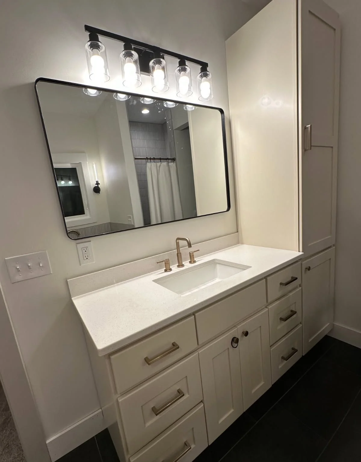 Modern bathroom vanity with white countertop, gold fixtures, large mirror, and cabinet storage, illuminated by four light bulbs.