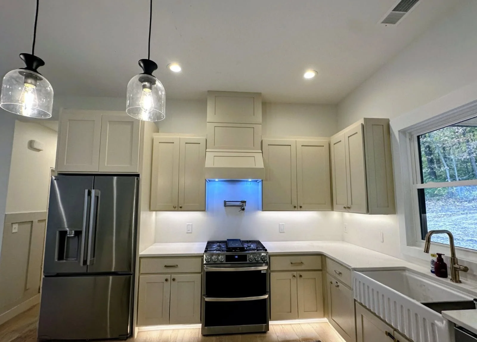 Modern kitchen with beige cabinets, stainless steel refrigerator, stove, and window over the sink, illuminated by ceiling and pendant lights.