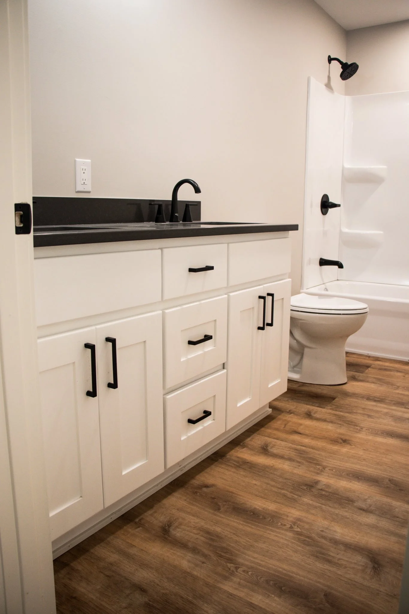 Bathroom with white vanity, black handles, black countertop, black faucet, beige wall, white toilet, shower with black fixtures, and wood-look flooring.