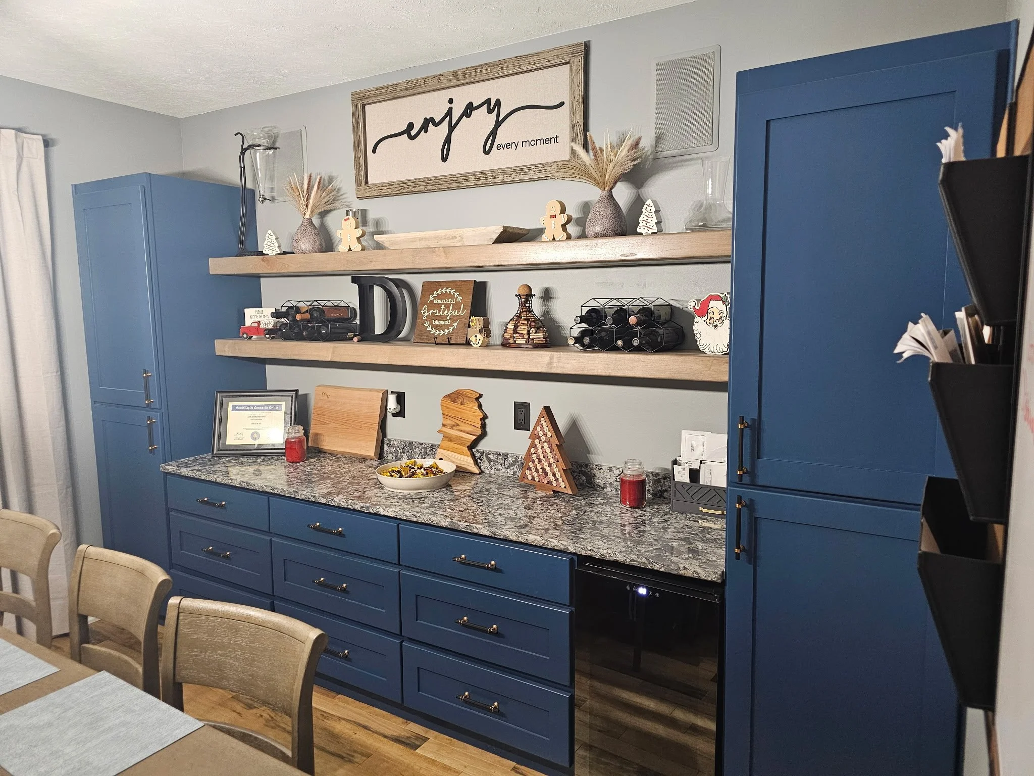 Blue kitchen cabinets with granite countertops, decorated for the holidays with small decor items, wine racks, and framed pictures, above a dining table with wooden chairs.