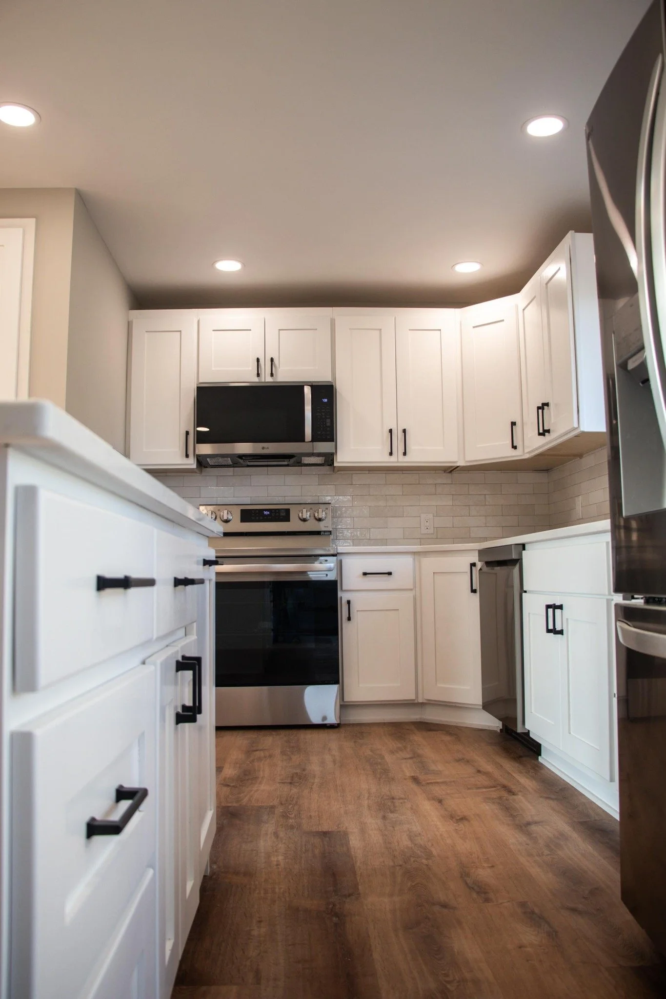 Modern kitchen with white cabinets, black handles, stainless steel appliances, wood flooring, and a beige brick backsplash.