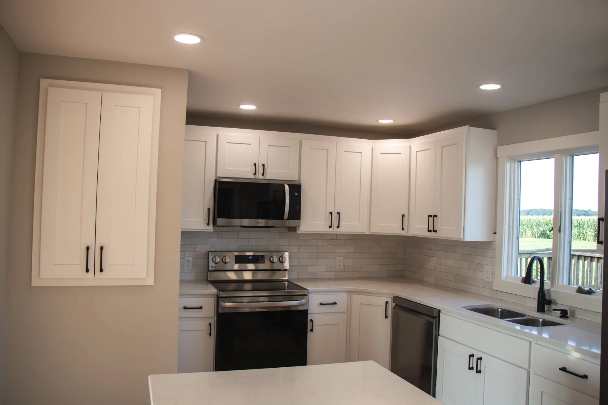 Modern kitchen with white cabinets, stainless steel appliances, a window showing a field, a black faucet, and recessed ceiling lights.