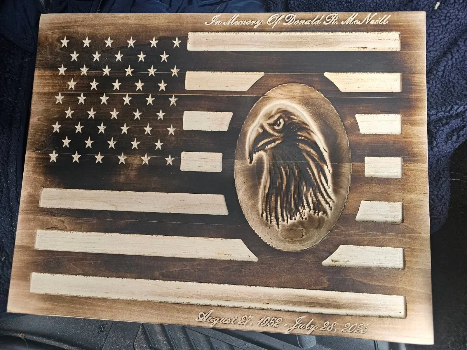 Wooden memorial plaque featuring an American flag with striped and star elements. The center has an engraved eagle's head. Text at the top reads 'In memory of Donald R. McNeill.' The bottom has dates 'August 21, 1959 - July 28, 2023.'