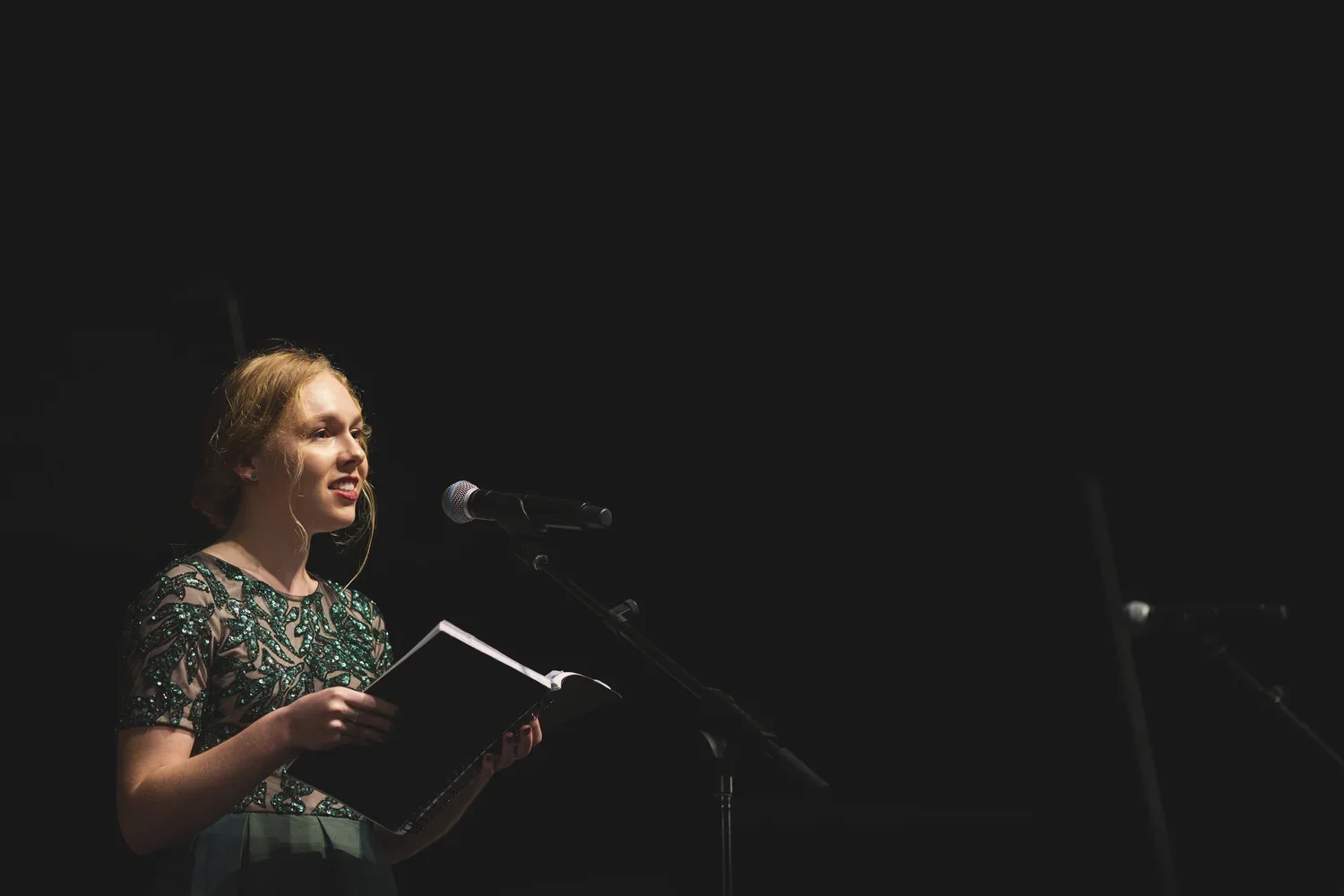 Young woman with wavy blonde hair, wearing a dress with green embellishments, stands at a microphone on a dark stage, holding a script or book, possibly performing or speaking.