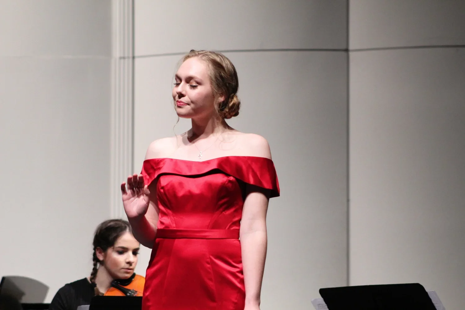 A woman in a red off-shoulder dress performing, with a woman in the background playing a musical instrument.