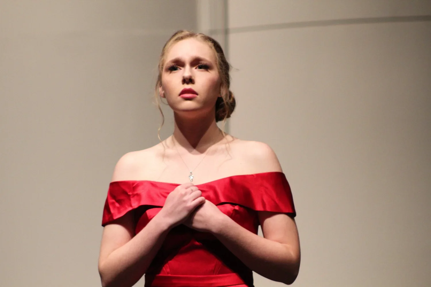 Young woman in a red off-shoulder dress with hands over her chest, looking up with a serious expression against a plain background.