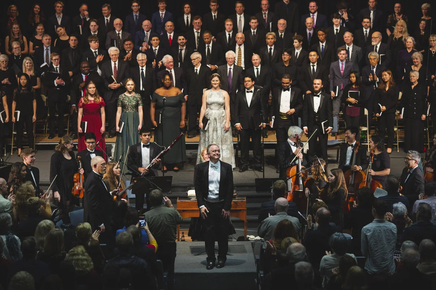 A large choir and orchestra performing on stage after a concert, with a conductor in front and audience clapping.