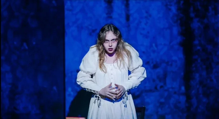 Young woman with long wavy hair wearing a white dress with puffed sleeves, standing with hands clasped in front, in a dark blue room or stage setting.