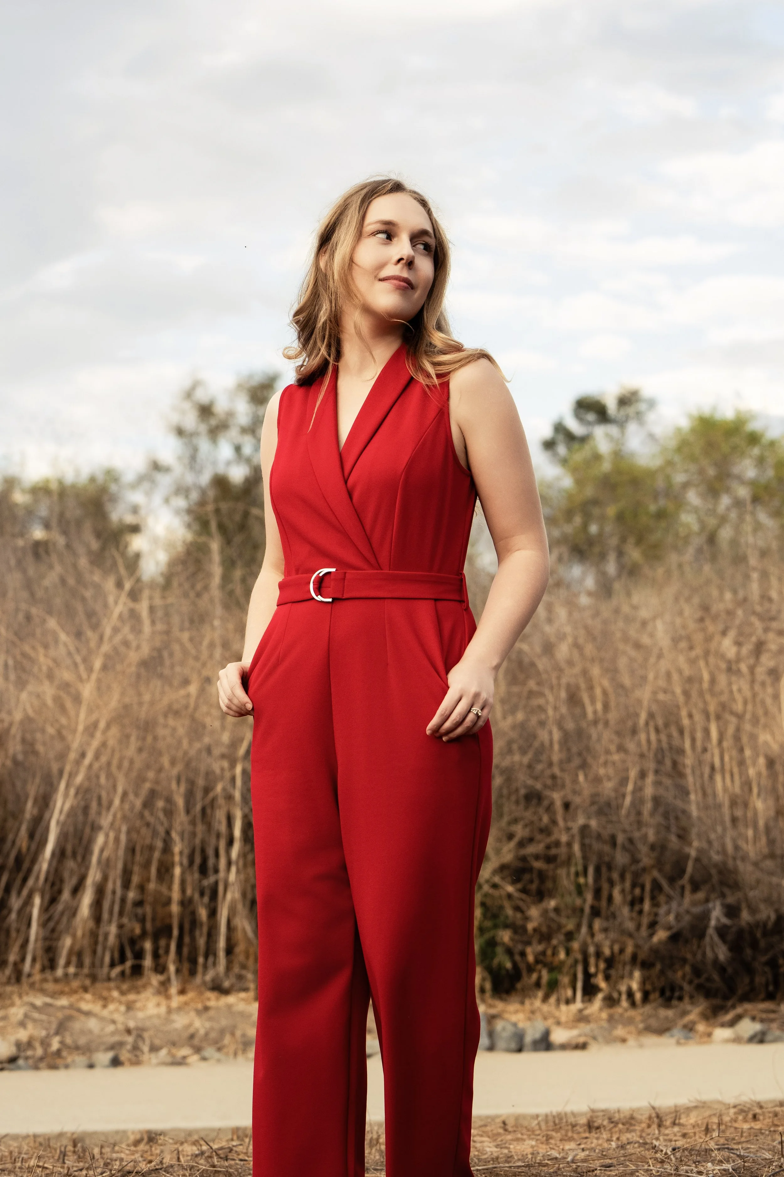 Woman in a sleeveless red jumpsuit standing outdoors with a background of bare trees and a partly cloudy sky.