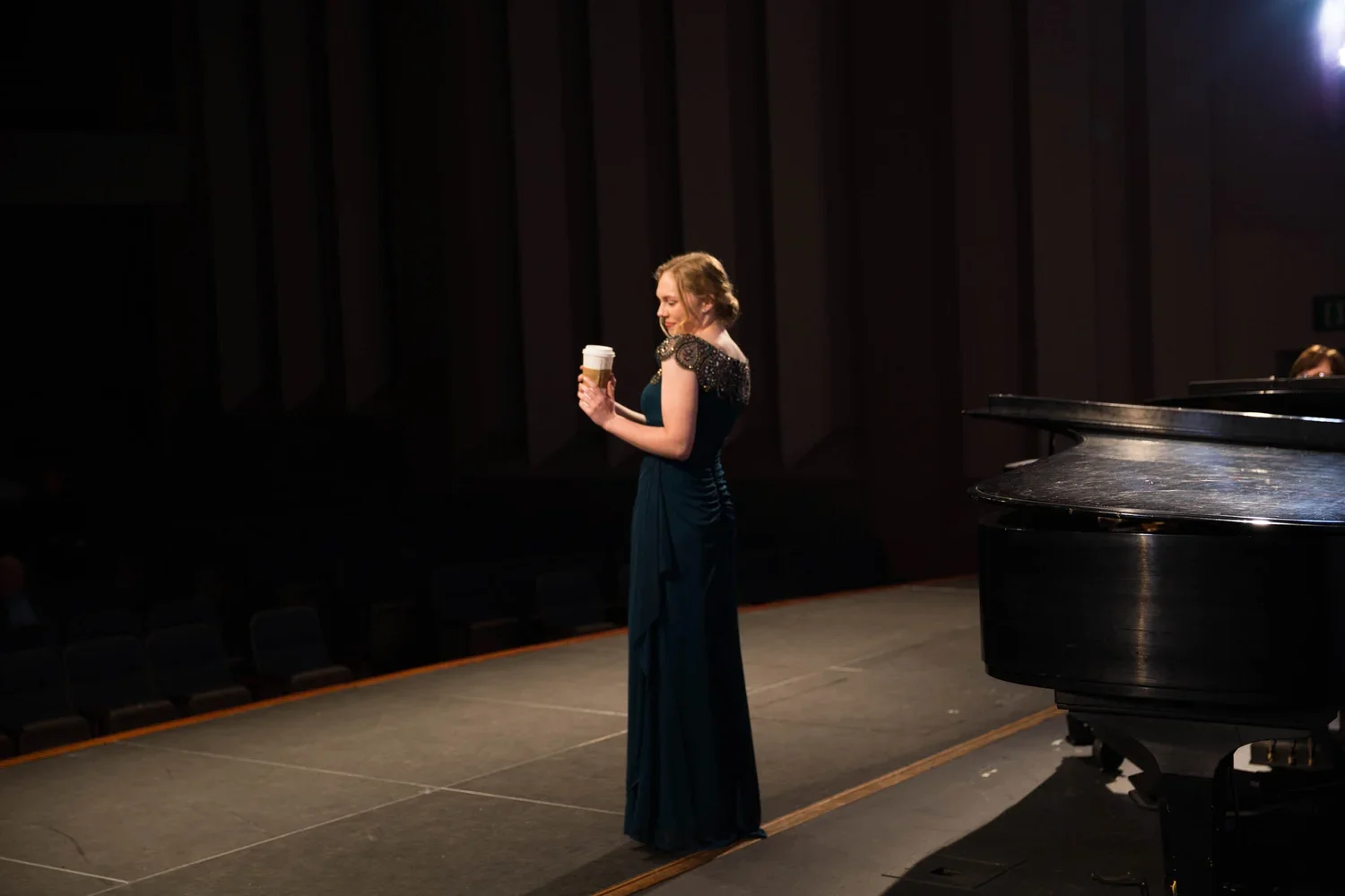 A woman in a formal dark gown on stage, holding a cup of coffee, standing near a grand piano, in a dimly lit theater or concert hall.