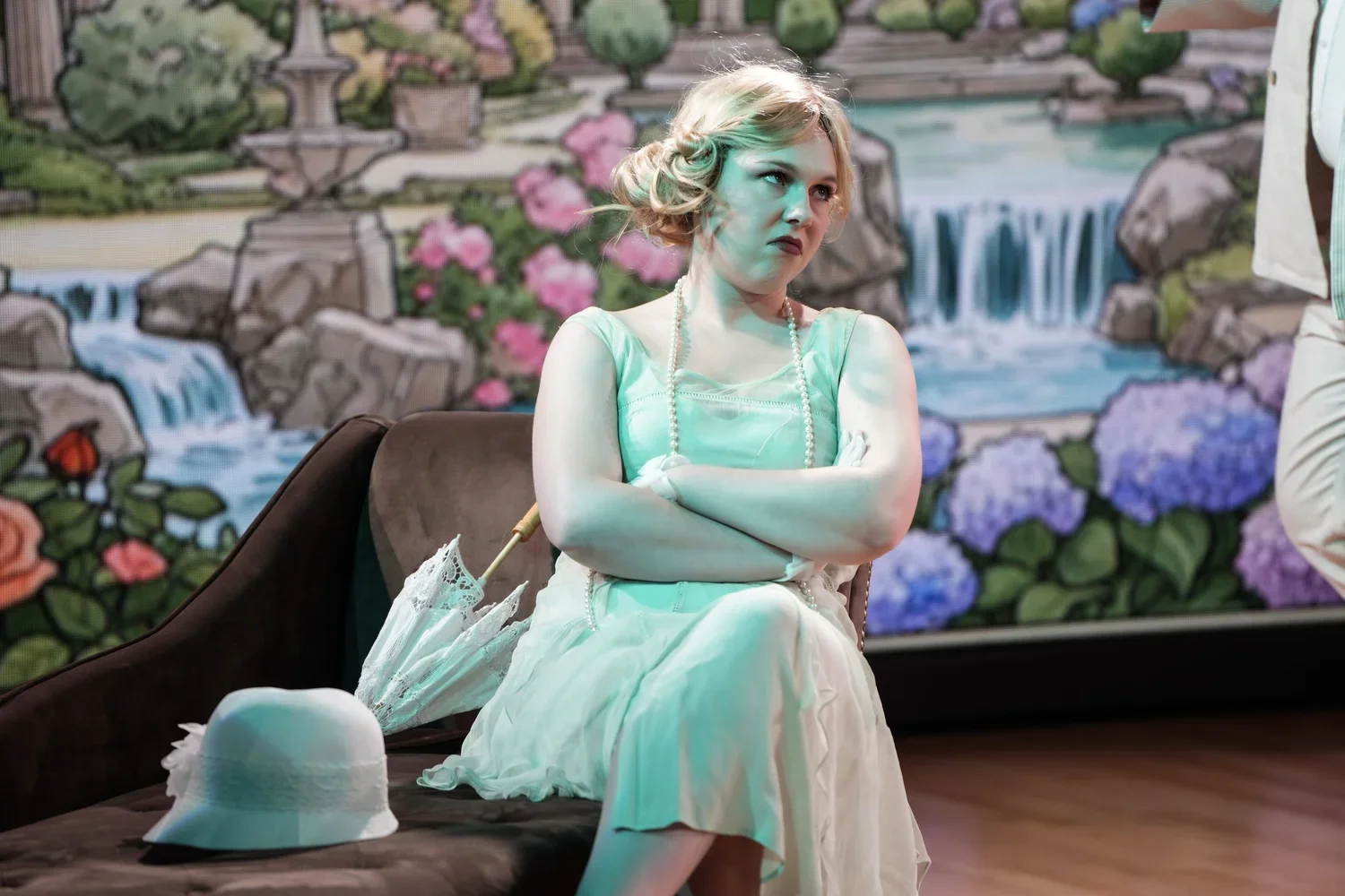 A woman with blond hair sits with crossed arms on a brown sofa. She wears a light green dress, a pearl necklace, and has a disapproving expression. There is a white hat and a lace umbrella beside her on the sofa. The background is a painted mural of a garden with flowers and waterfalls.
