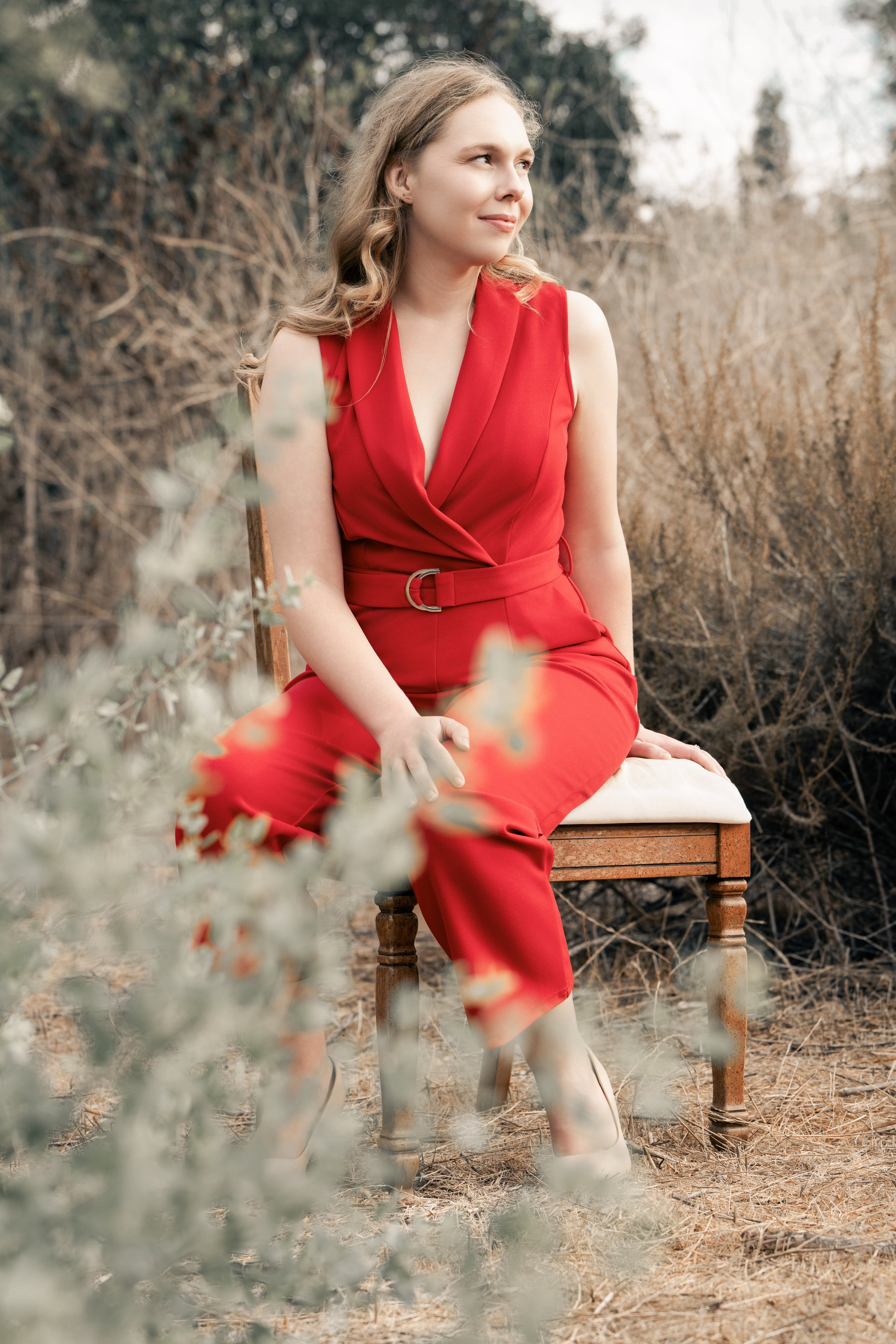 A young woman in a red sleeveless dress sitting on a wooden chair outdoors, with mountains and trees in the background.