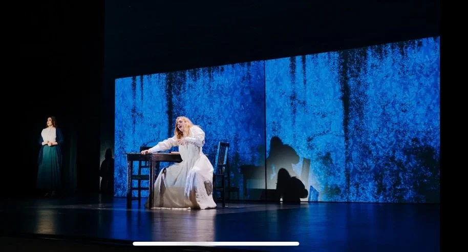 A woman in a white dress sitting at a table with a keyboard, with a woman in dark clothing standing to the left and blue textured background on stage.