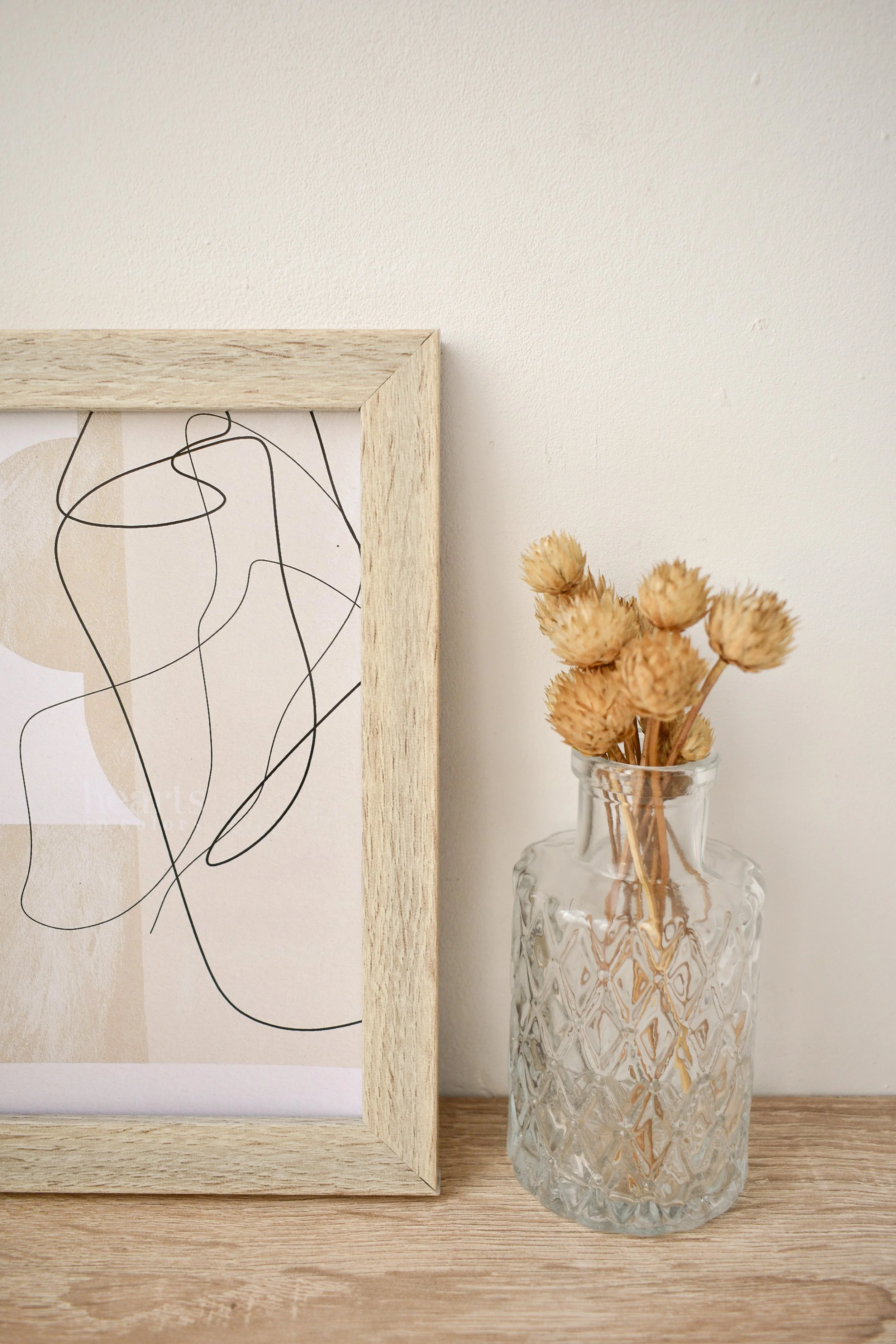 A glass vase with textured diamond pattern containing dried beige flowers, placed on a wooden surface next to a framed abstract art print with black lines on a beige background.