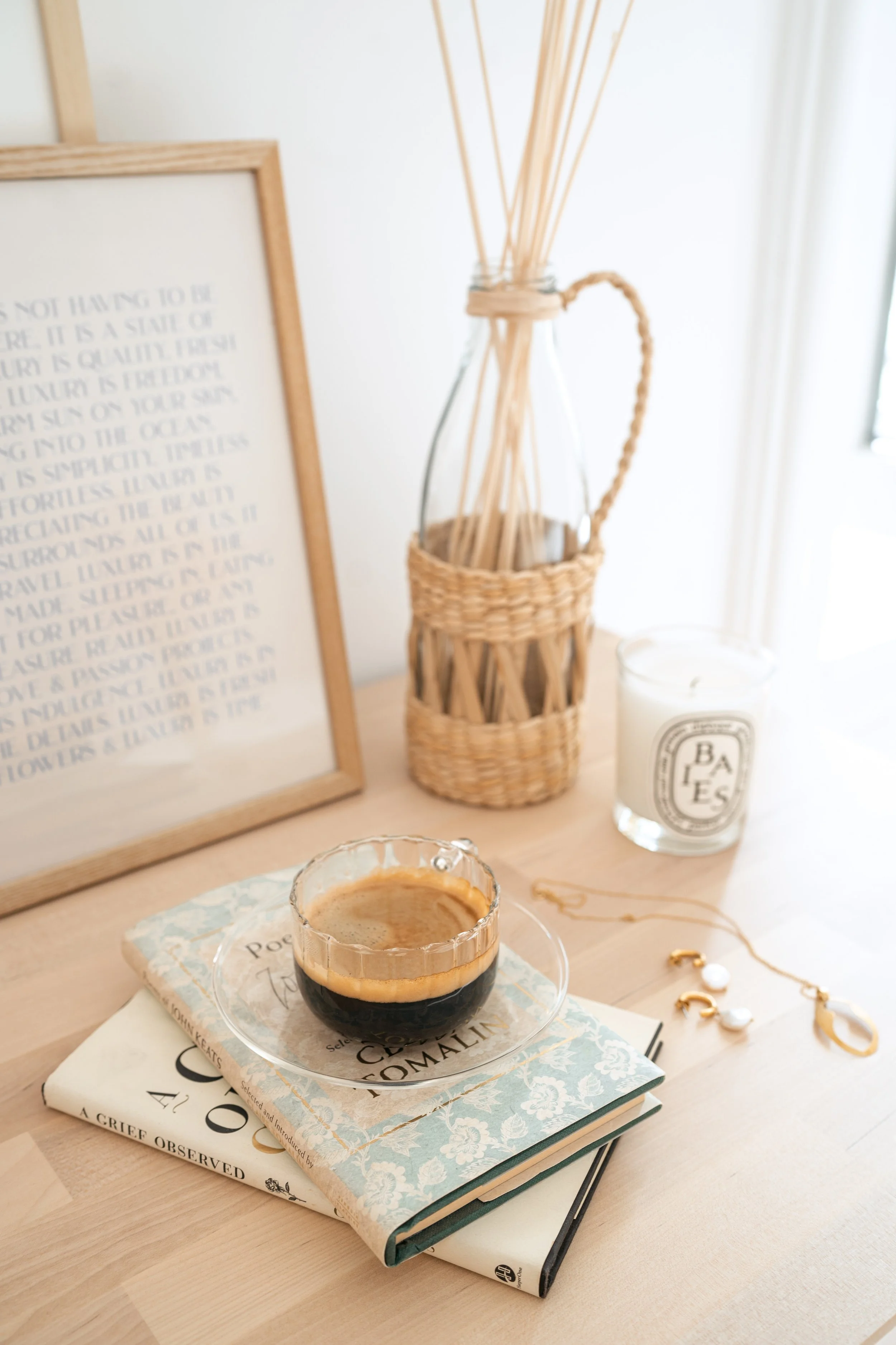 A cup of black coffee on a stack of books on a wooden table, with a framed text, a reed diffuser, a white candle, and jewelry in a well-lit room.