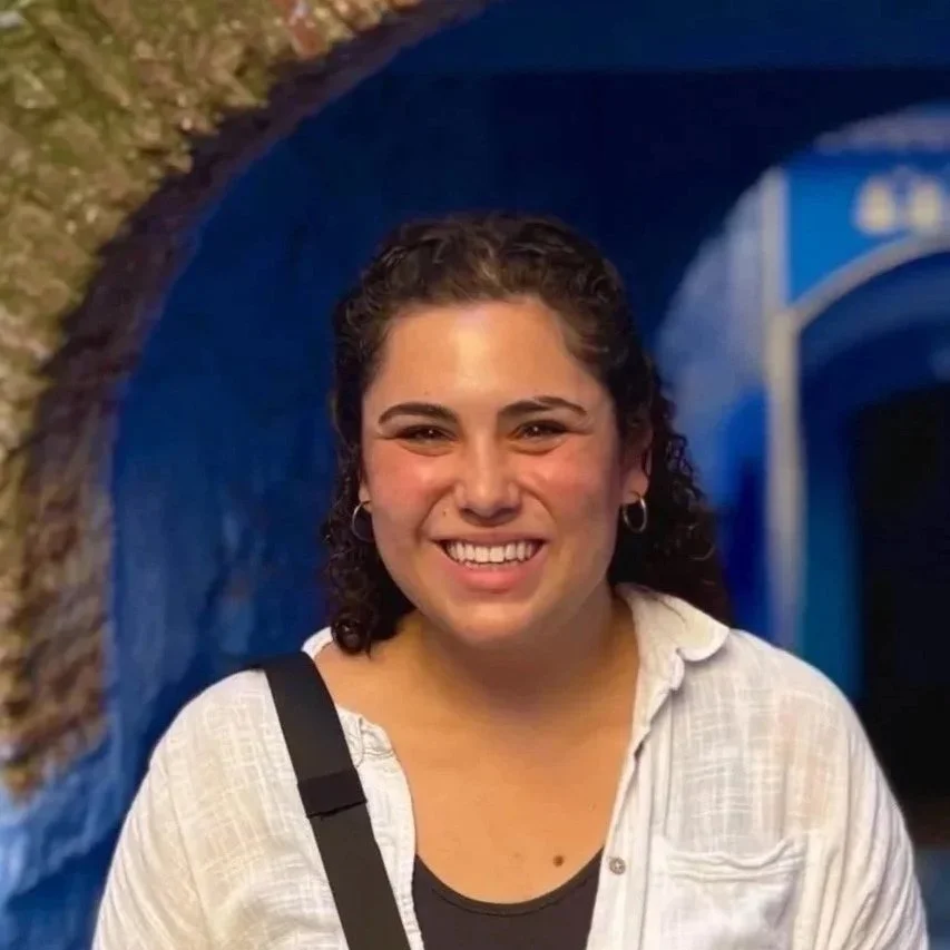 A woman with dark, curly hair smiling outdoors in a blue-painted market