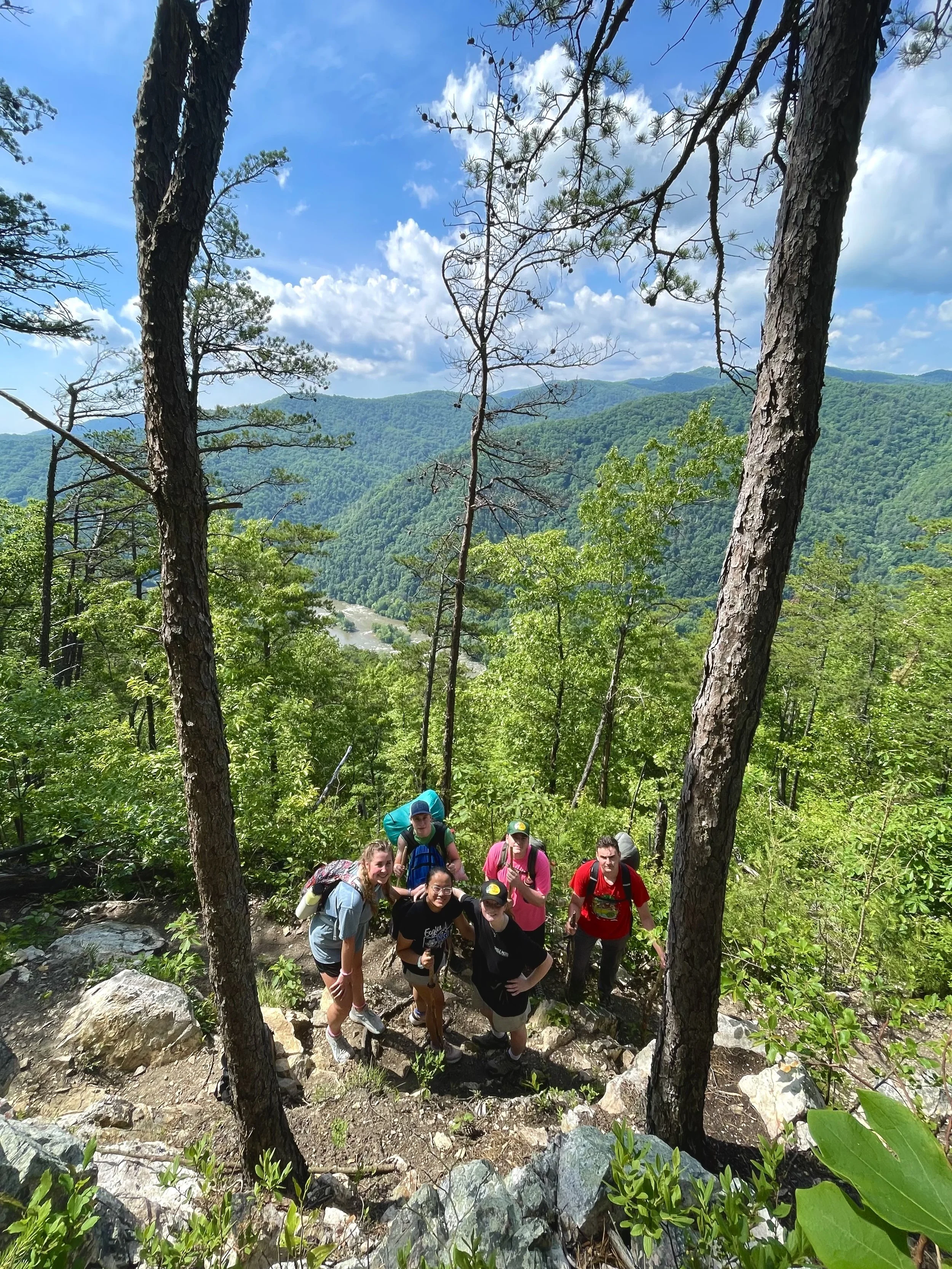 Group of hikers climbing a rocky trail on a lush green mountain with blue sky and clouds above.