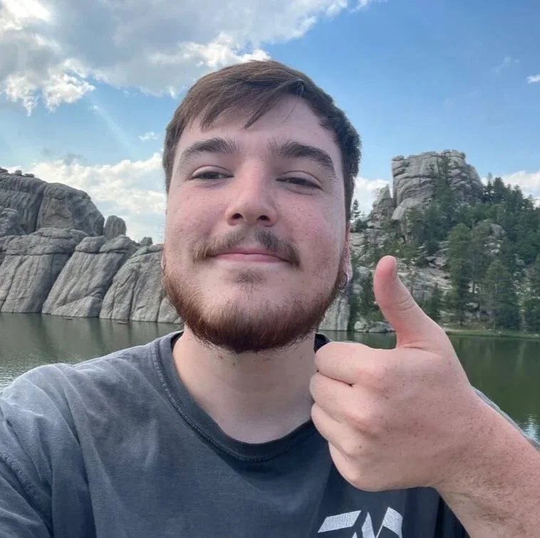 A young man with brown hair and a beard giving a thumbs-up gesture outdoors, with a lake, rocky cliffs, and a partly cloudy sky in the background.