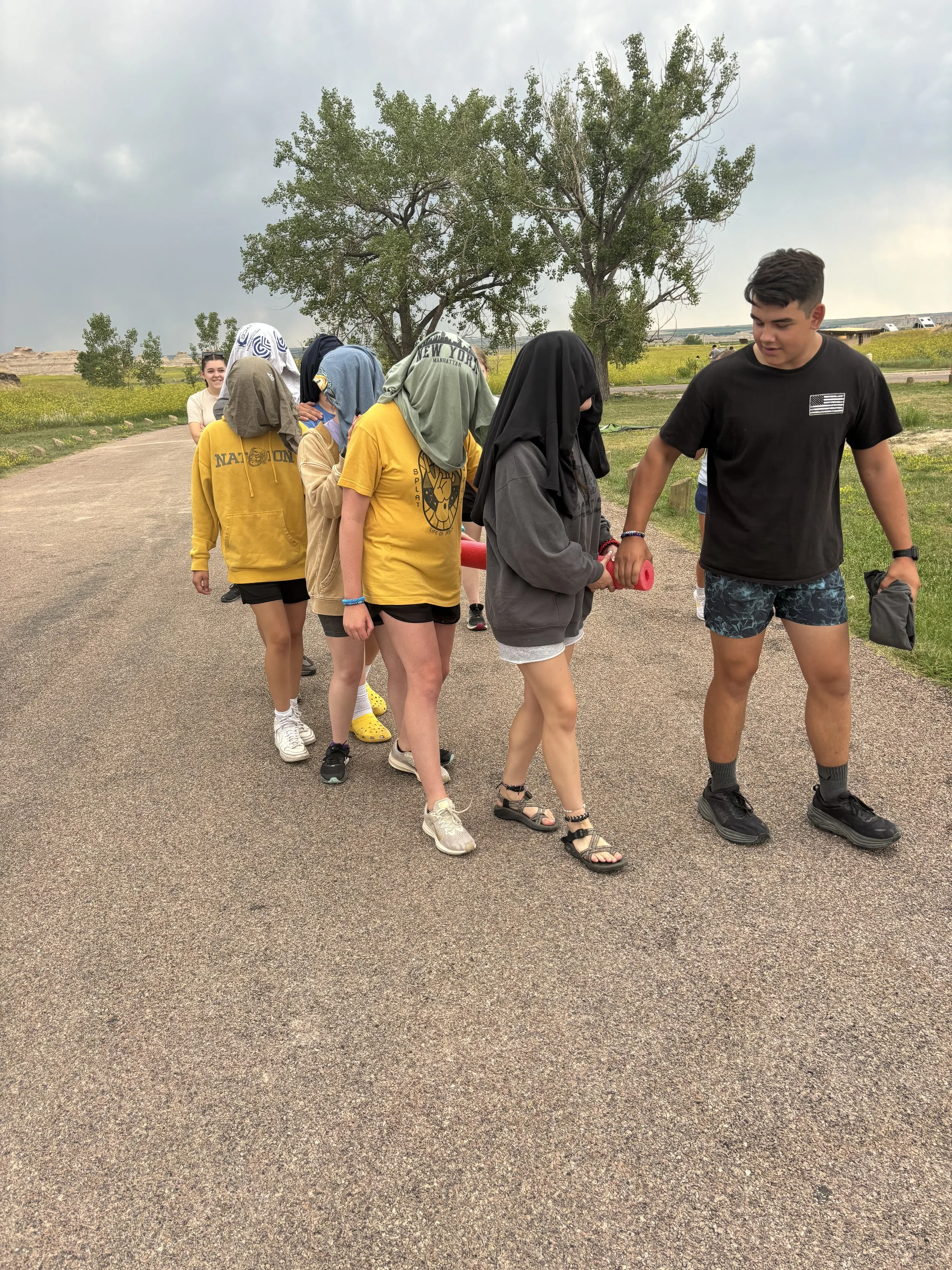 A group of children and teenagers outdoors on a pathway, some with their heads covered by shirts or hoodies, and one person helping another with a foam roller, with trees and open fields in the background on a cloudy day.