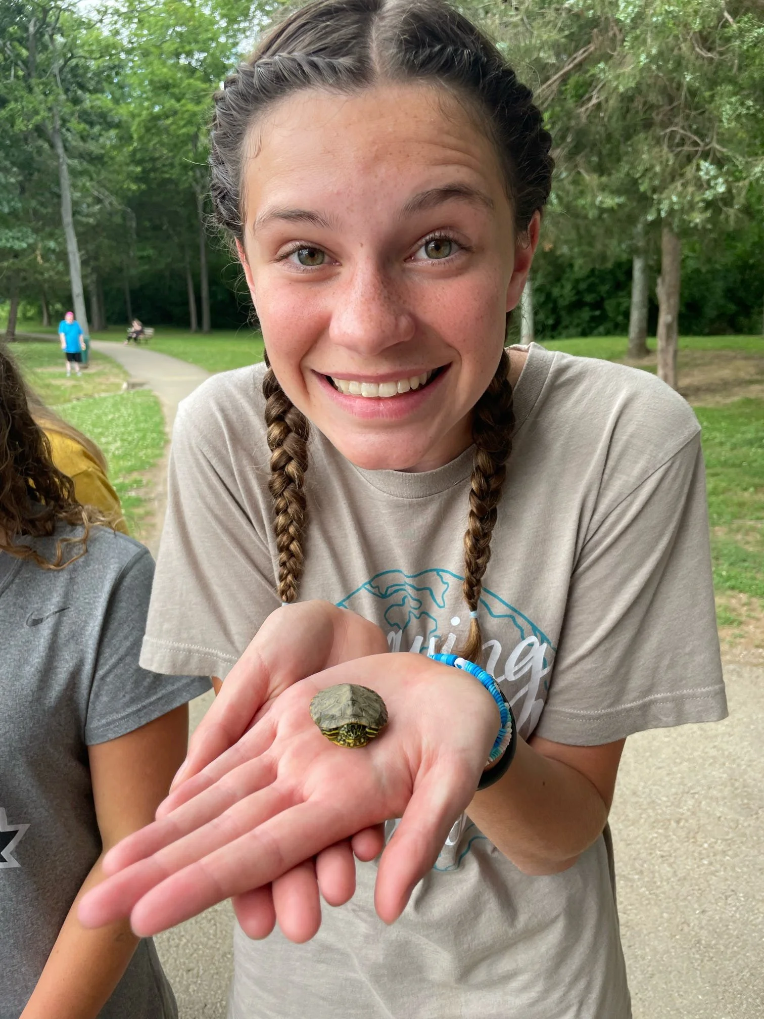 Young girl with braided hair smiling and holding a small turtle on her palm in a park.