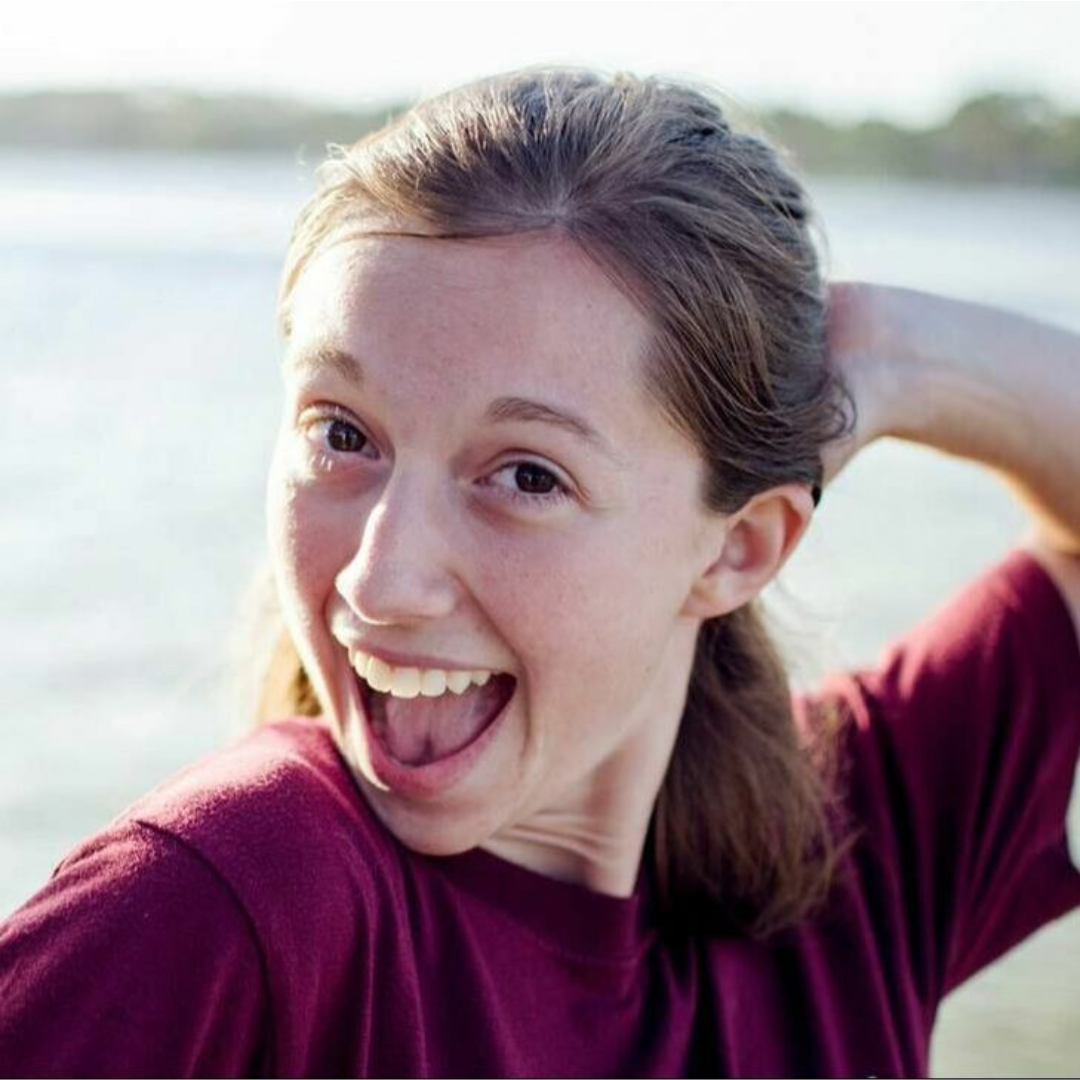 A young woman with brown hair, smiling and showing her teeth, wearing a maroon shirt outdoors near water with a blurred background.