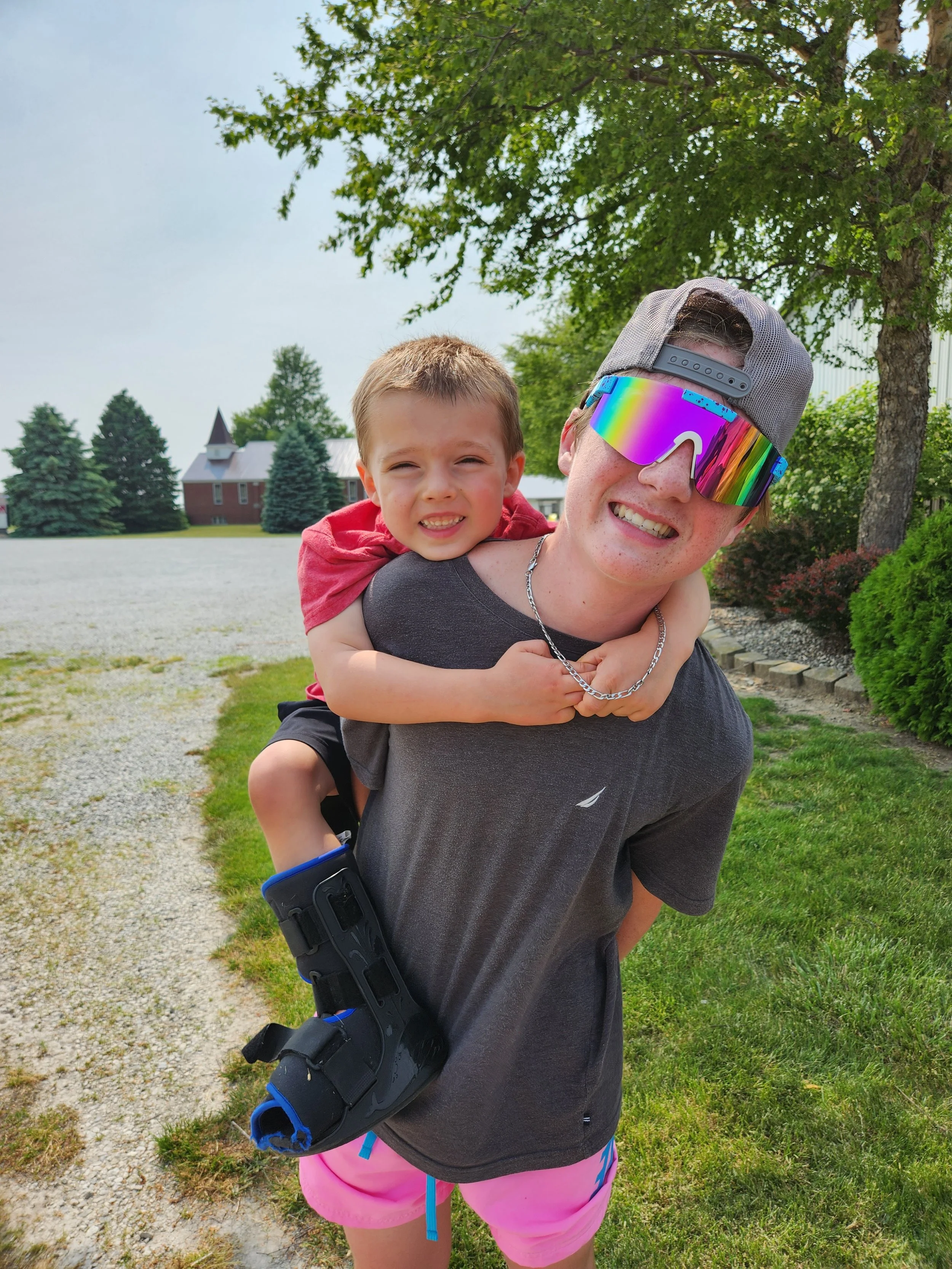 A boy with a red shirt riding on the shoulders of a teenager with sunglasses and a gray cap, smiling outdoors on a sunny day near houses and green trees.