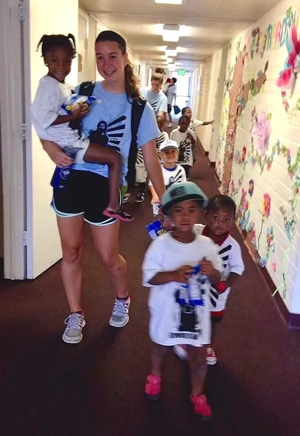 A group of children and a young woman walking down a hallway decorated with colorful artwork. The woman is carrying a girl in her arms, and the children are holding small items, possibly toys or snacks.