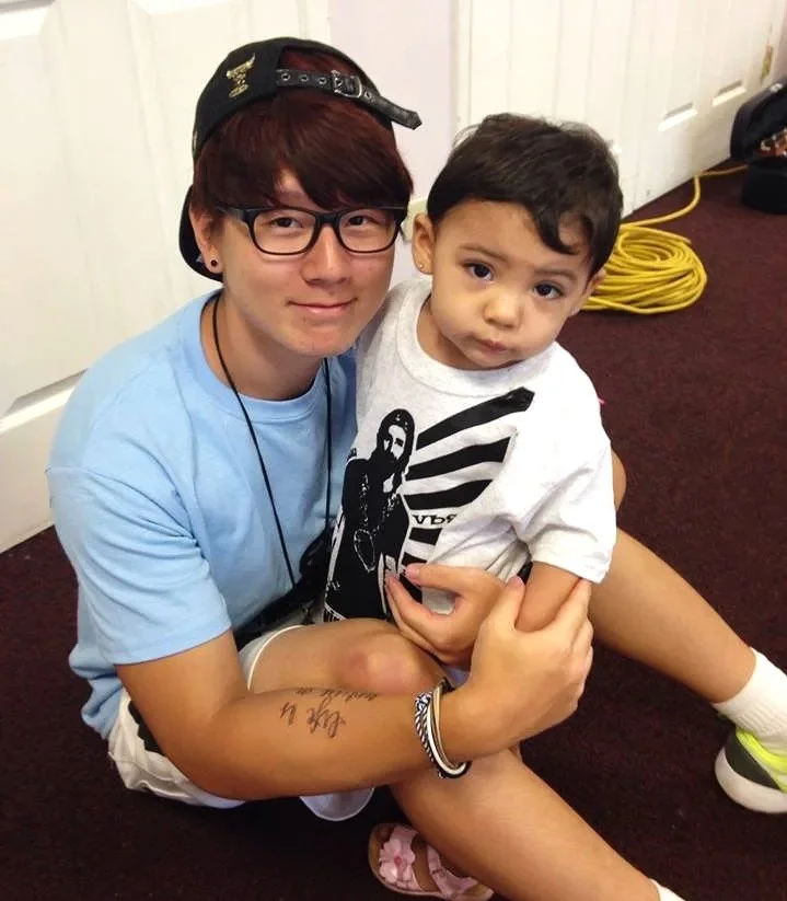 A young person with red hair, glasses, and a black cap sitting on the floor, holding a small boy with dark hair and a white T-shirt featuring a graphic design. The young person is smiling, and the boy has a neutral expression.