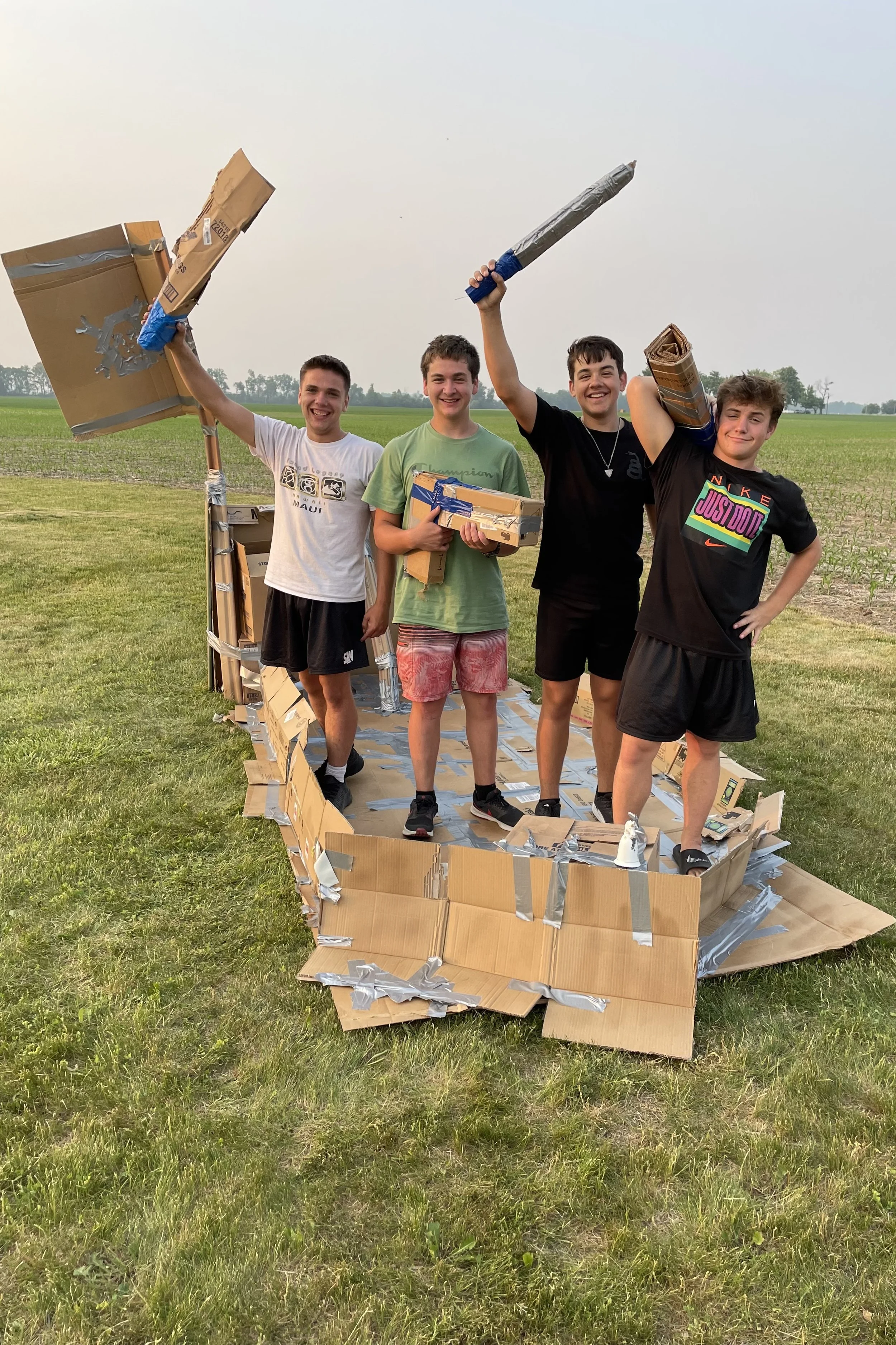 Four young boys standing inside a large handmade cardboard rocket ship with metallic tape details, celebrating outdoors in a grassy open field, holding what appear to be makeshift rocket props.