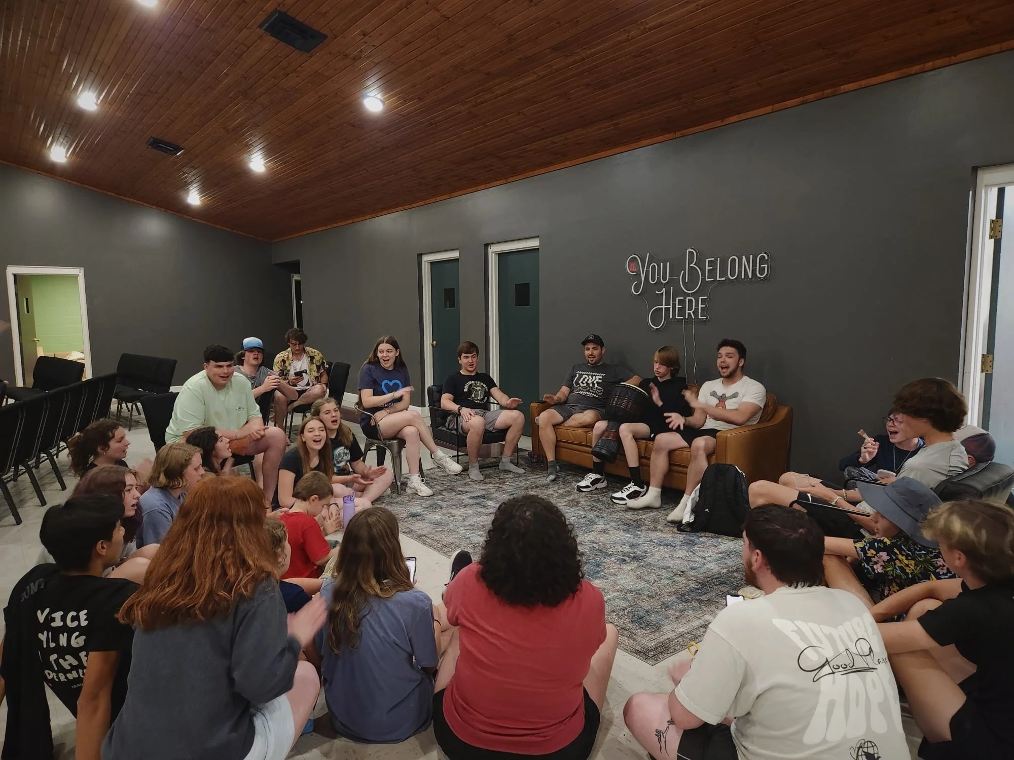 Group of teenagers and young adults seated in a circle in a room with dark gray walls, wooden ceiling, and neon sign saying 'You Belong Here'.