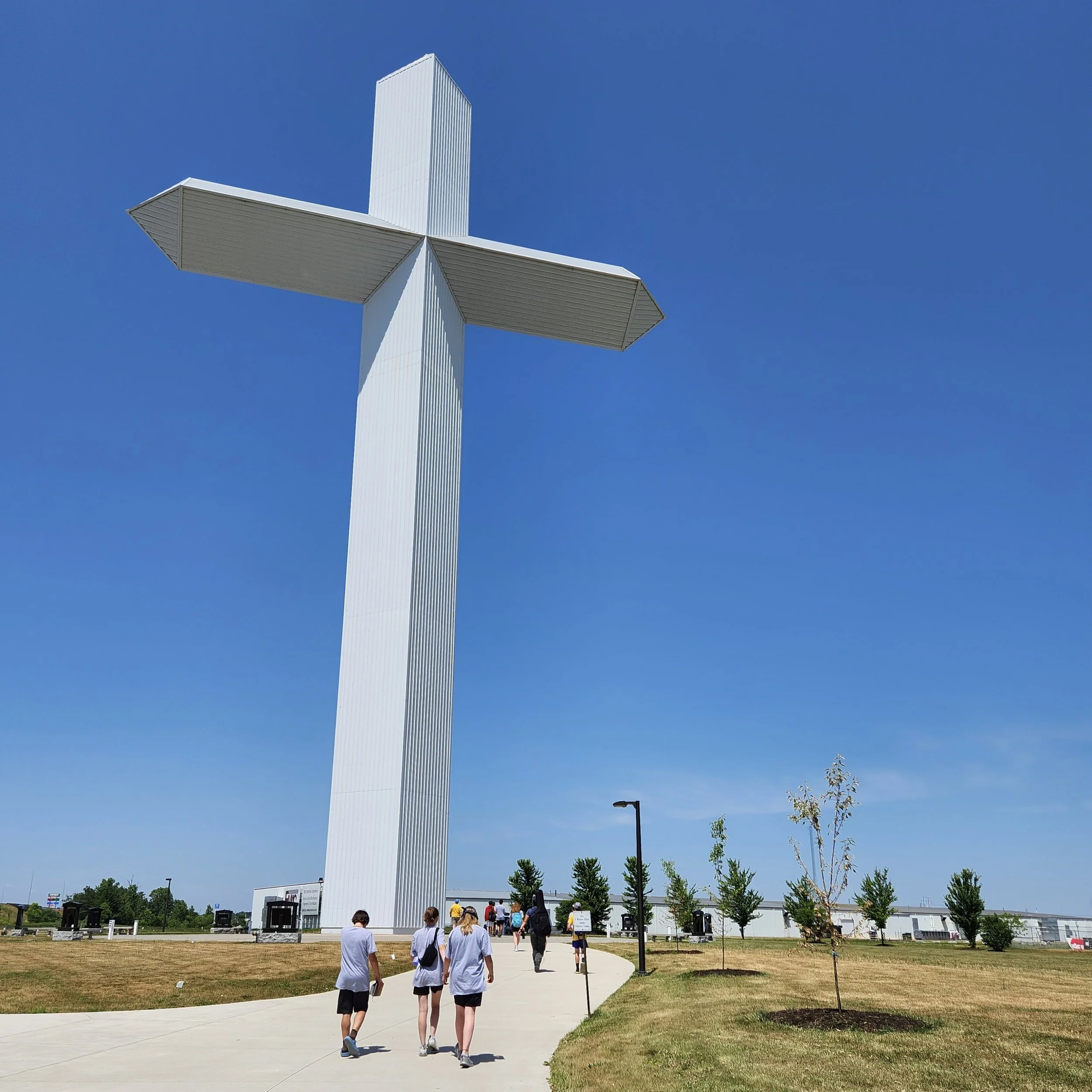 A large white cross monument with a modern design, situated outdoors under a clear blue sky, with several people walking along a paved path toward it, surrounded by a grassy area and small trees.