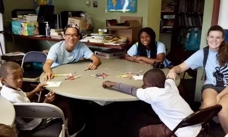 Four children sitting around a table with one child reaching out, others smiling and engaging in a group activity or game in a classroom setting.