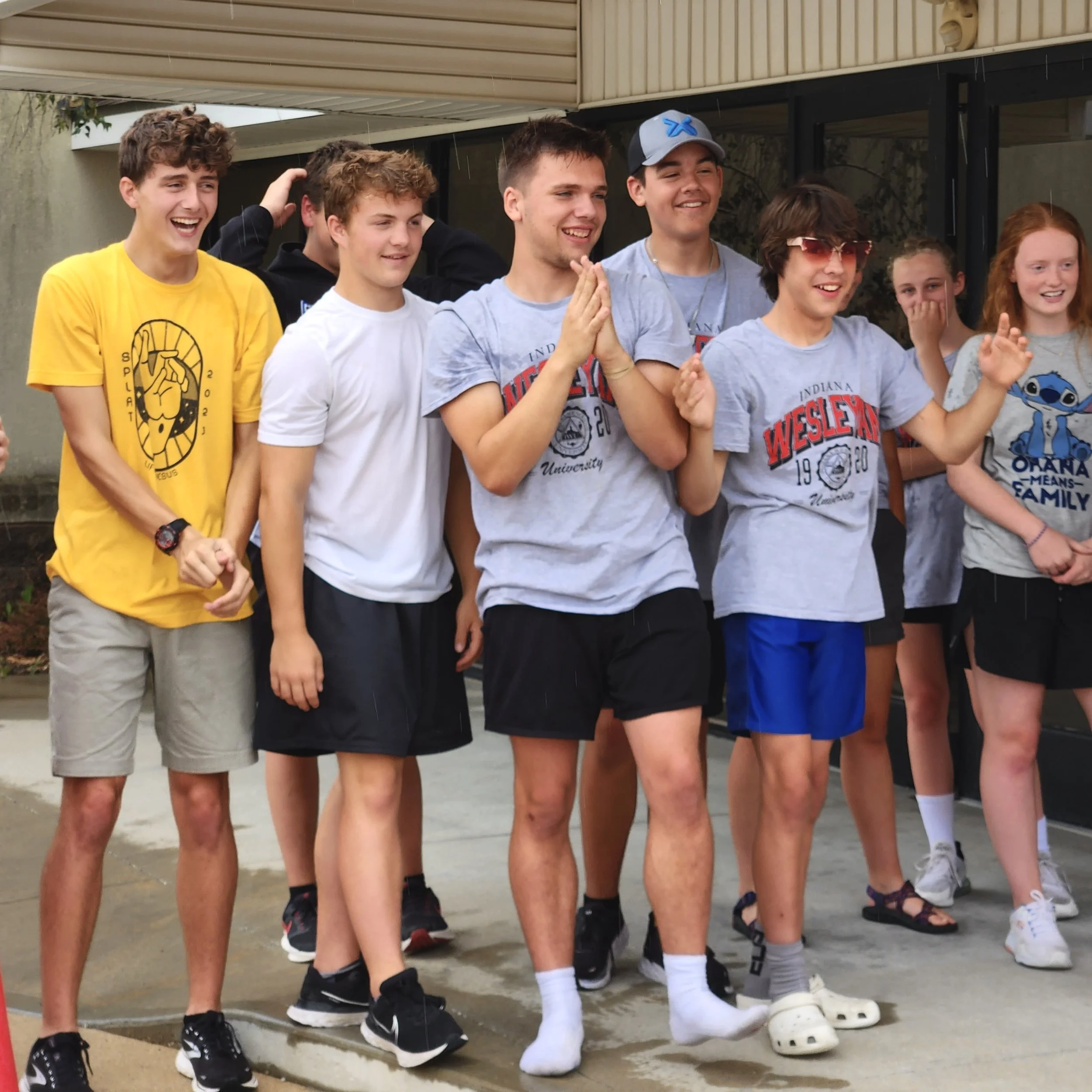 Group of teenagers standing outdoors, some clapping and smiling, dressed casually in T-shirts and shorts.