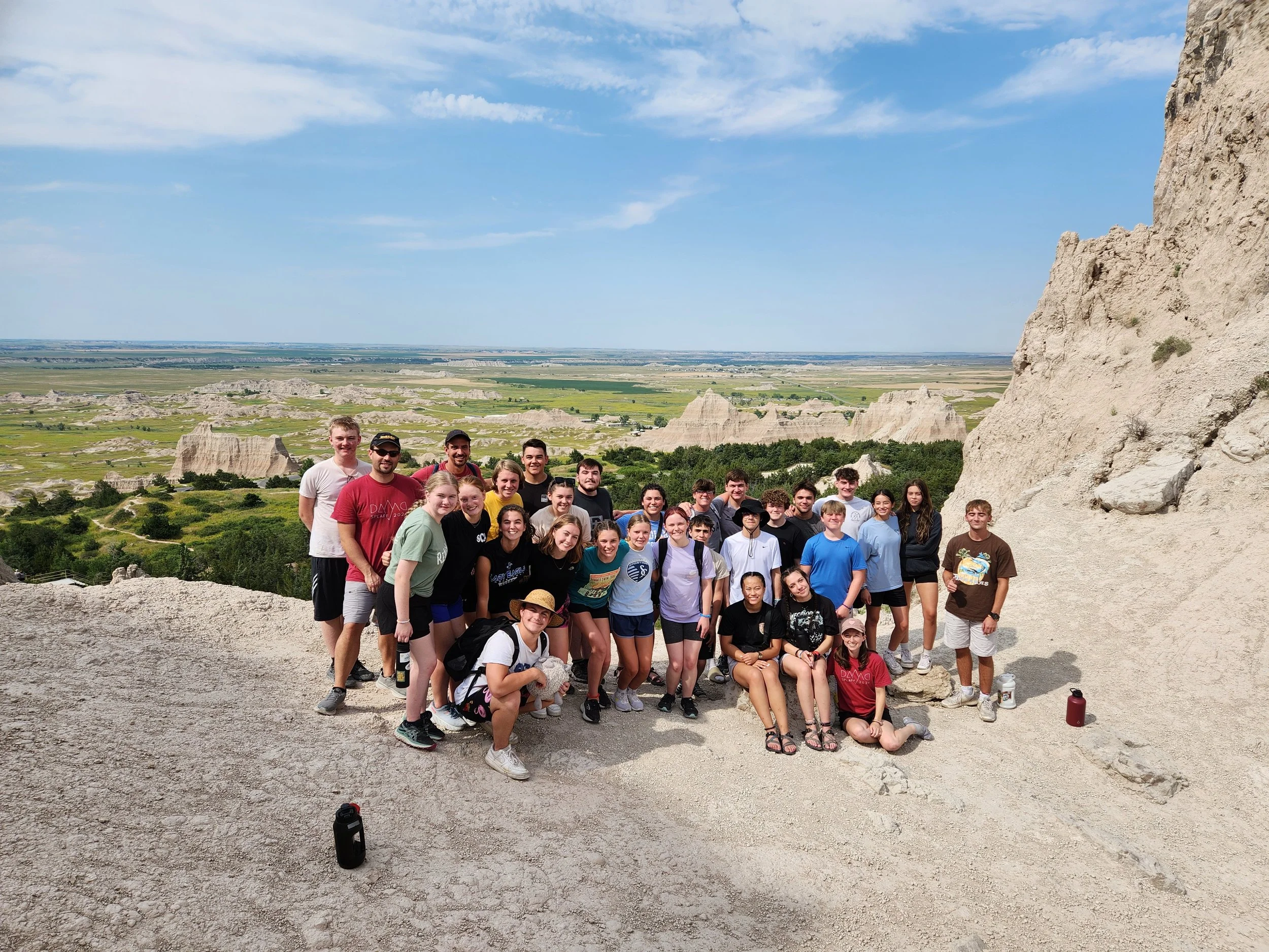 Group of young people and adults hiking on a rocky trail with a scenic view of a grassy valley and white rock formations in the background under a partly cloudy sky.