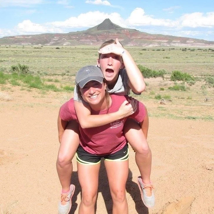 A woman giving a piggyback ride to a young boy in an open, desert-like area with a mountain in the background. Both are smiling and dressed casually for outdoor activity.