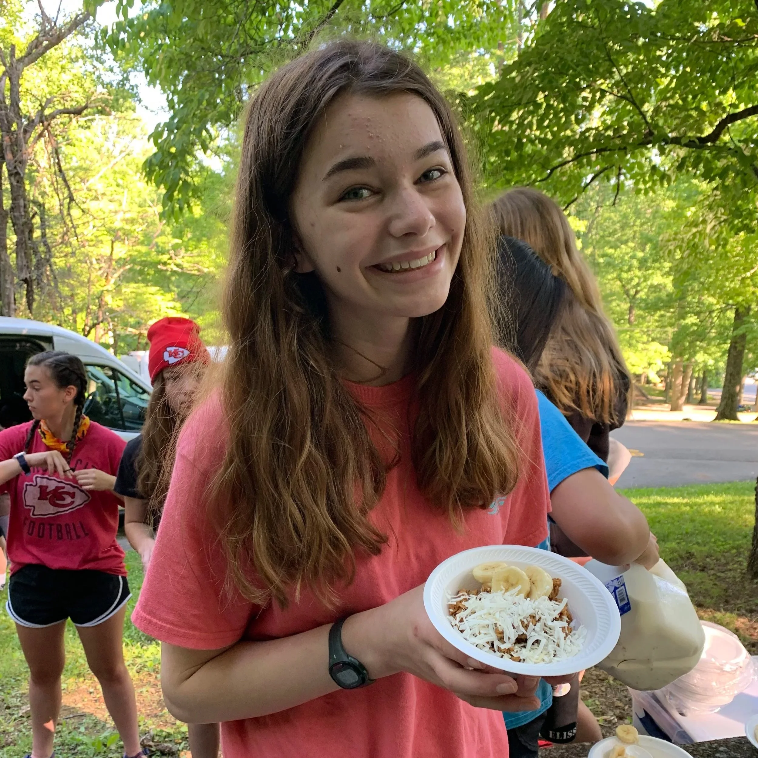 A young woman with long brown hair and light skin holding a small white plate of food, including sliced bananas and shredded cheese, smiling at the camera outdoors with green trees and a group of people in the background.