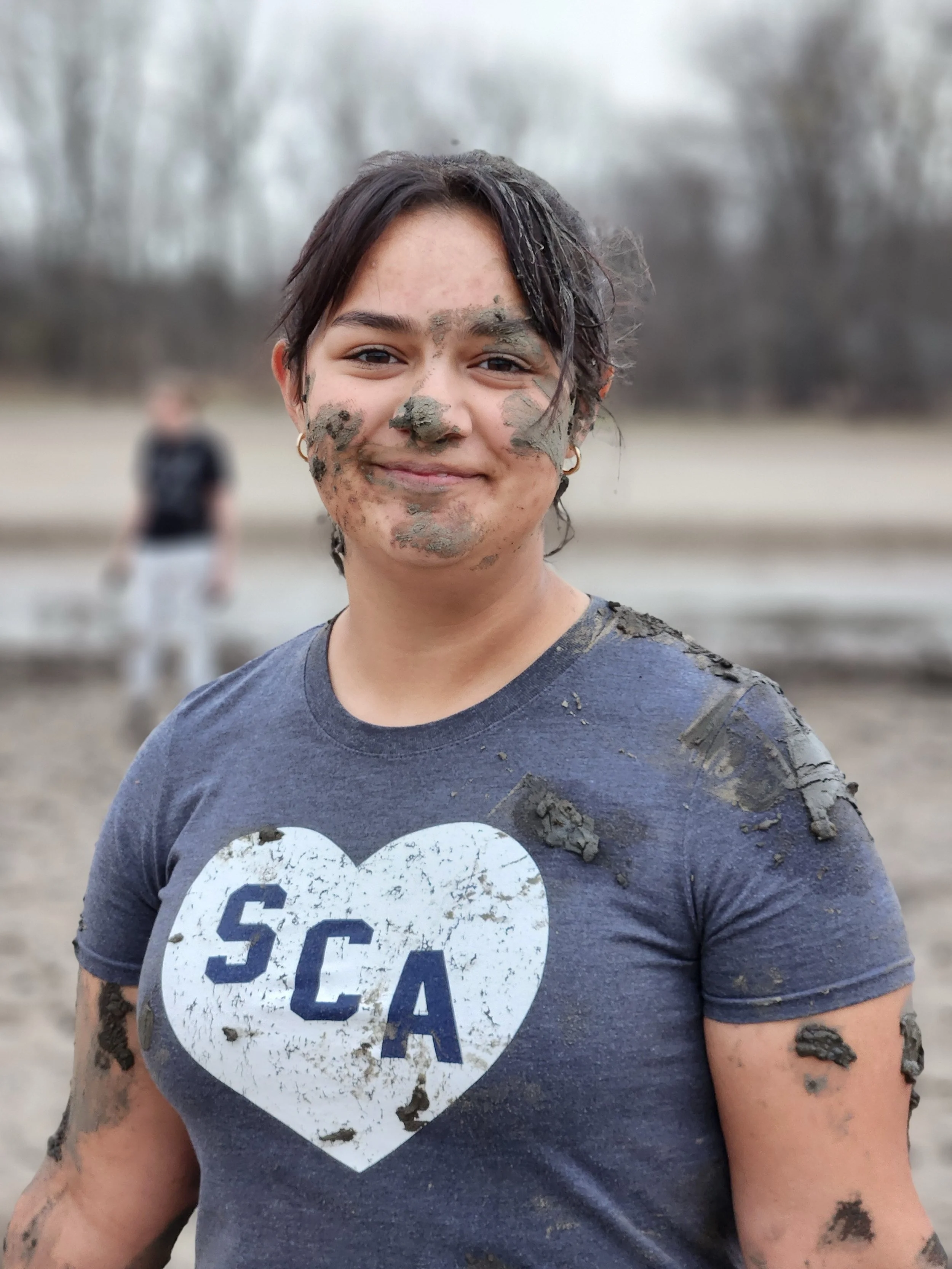Woman with mud on her face, arms, and t-shirt, smiling outdoors with a blurred background.