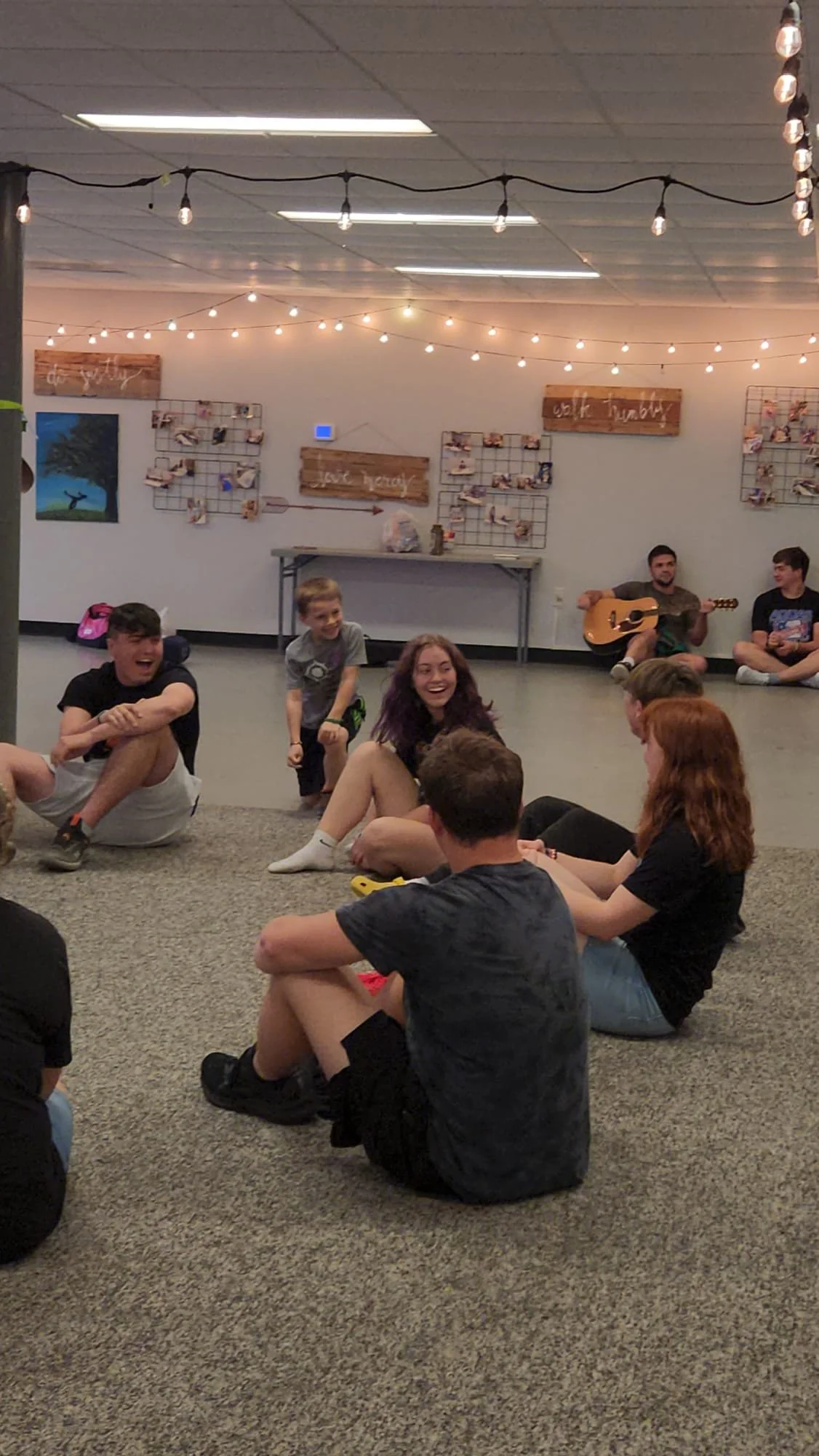 A group of people, mostly teenagers, sitting on the carpeted floor of a basement or large room, laughing and talking. In the background, some teenagers are sitting on the floor, one playing guitar and others chatting. The room is decorated with strin
