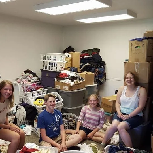 Four children sitting on the floor of a room with moving boxes and laundry baskets filled with clothes behind them, smiling at the camera.