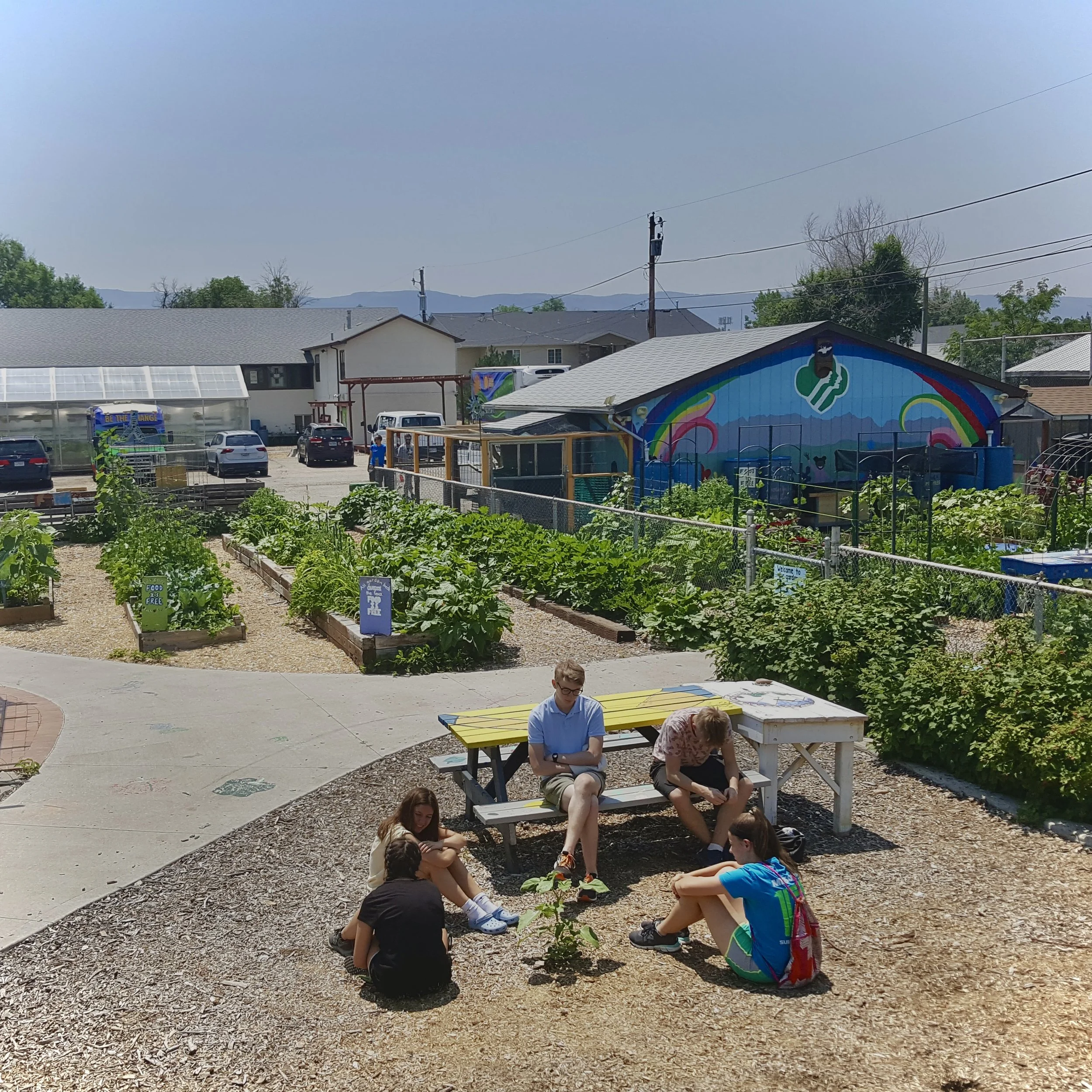 A group of five young people sitting on the ground and on a bench in a community garden with lush green plants, a colorful mural on a building, parked cars, and residential houses in the background.