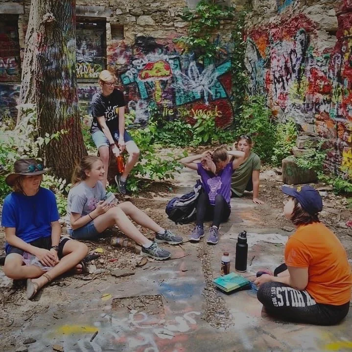 Group of five children sitting on the ground and one standing near a graffiti-covered brick wall and trees in an outdoor urban area. They appear to be engaged in a conversation or activity.