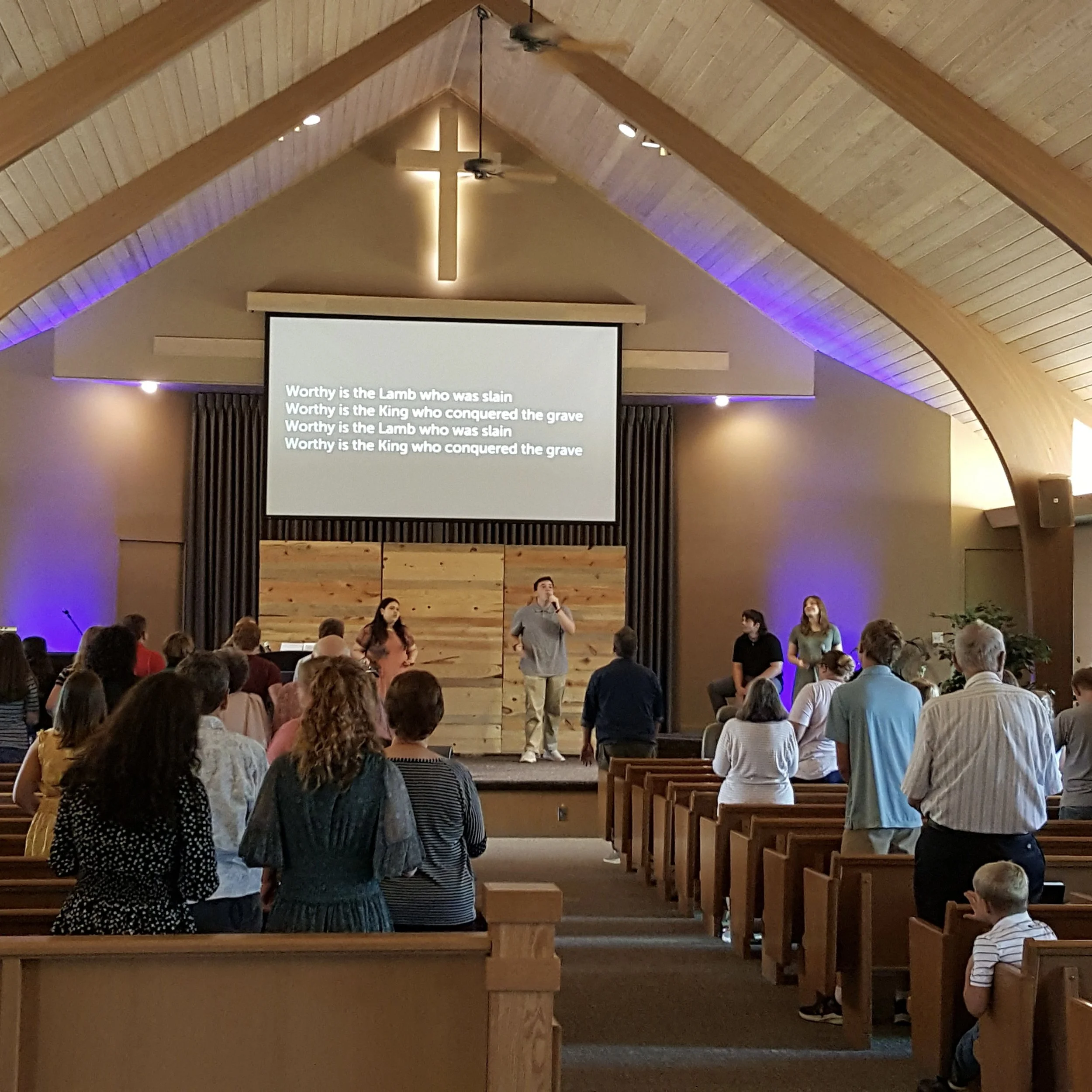A church service with people standing and singing in the pews, a worship leader on stage, a large cross illuminated behind the stage, and lyrics projected on a screen that read: 'Worthy is the Lamb who was slain, Worthy is the King who conquered the 